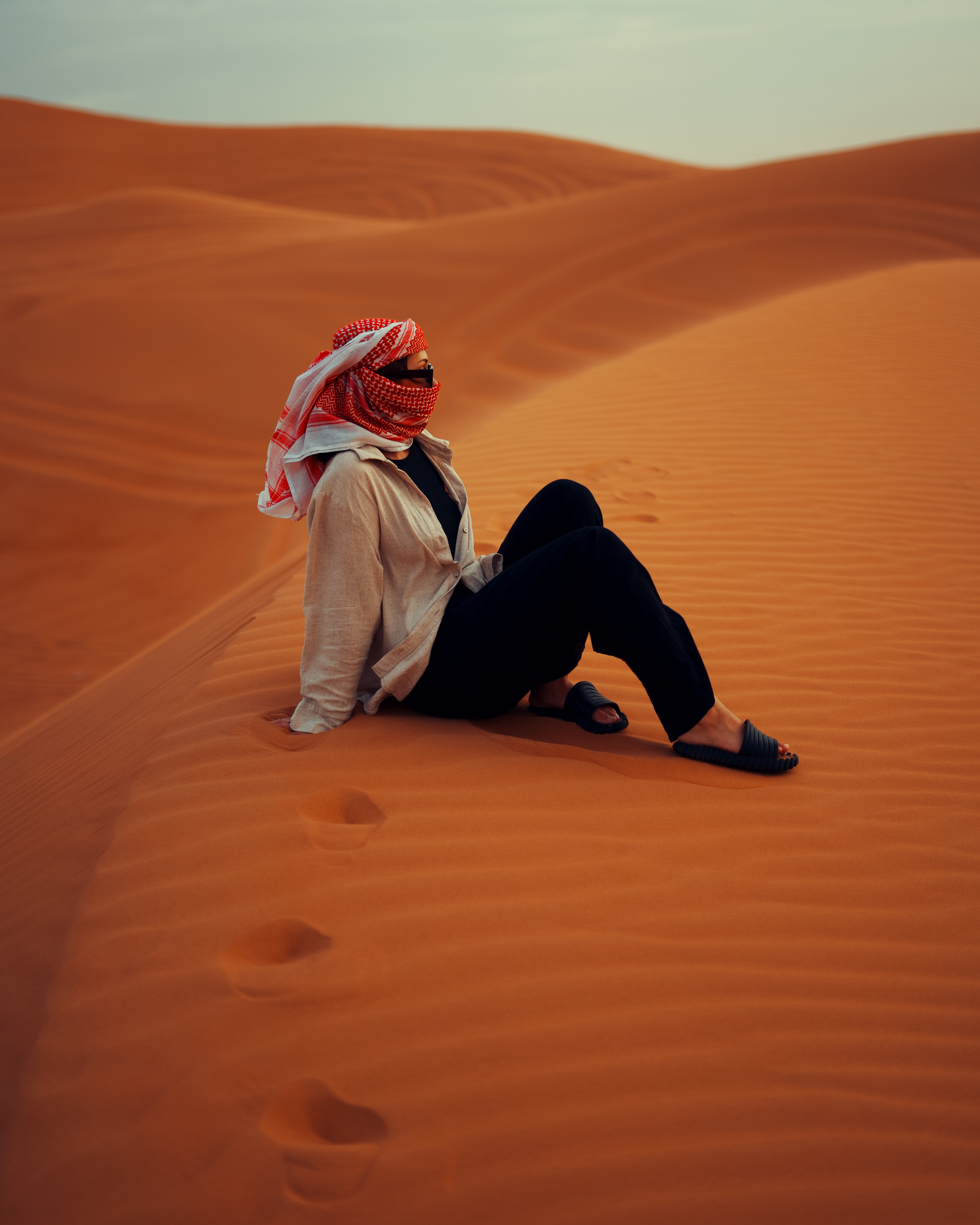 Person wearing a red and white headscarf, beige shirt, black pants, and sandals sitting on orange sand dunes with visible footprints.