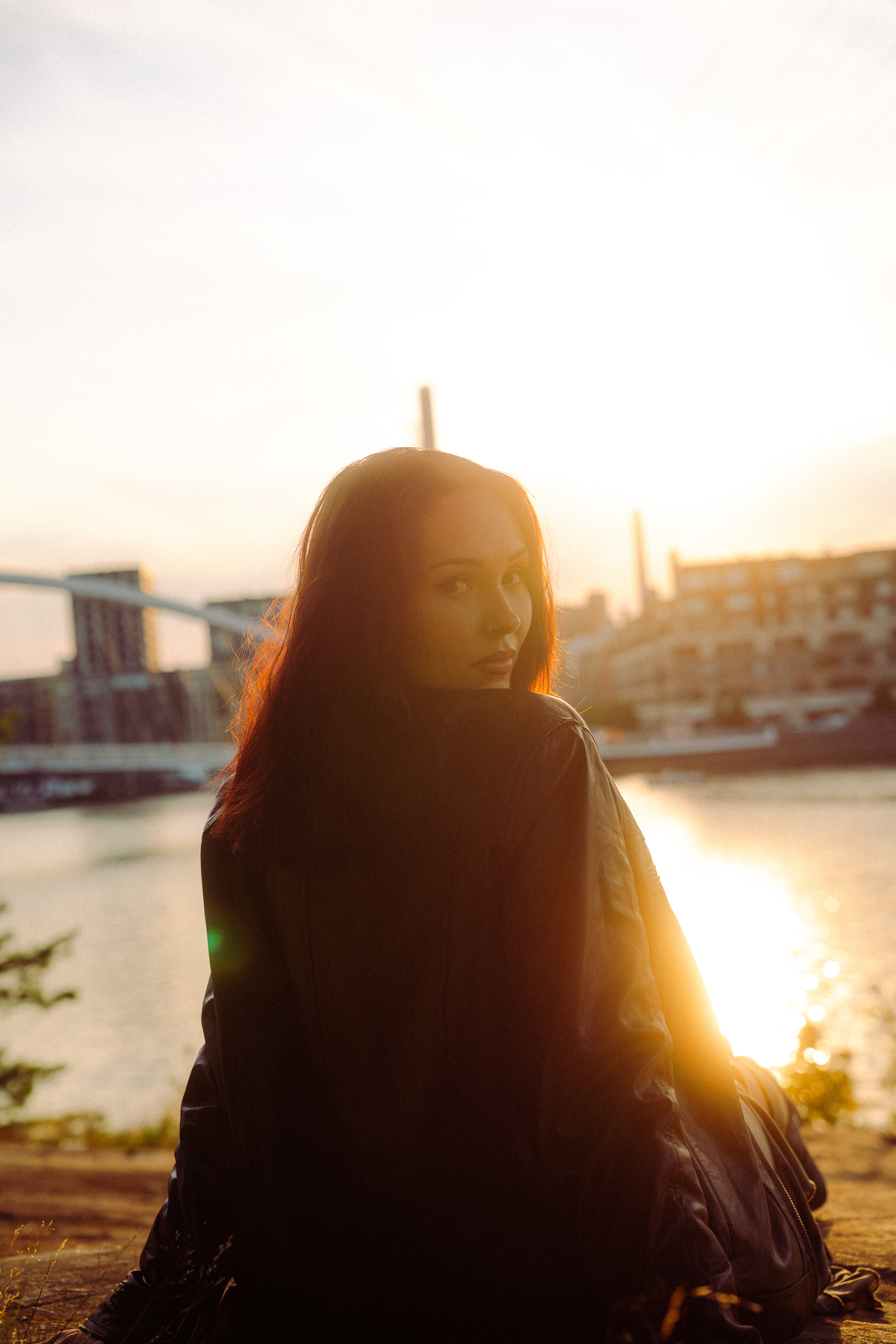 Young woman with long hair wearing a leather jacket sitting by a river at sunset, looking back over her shoulder.