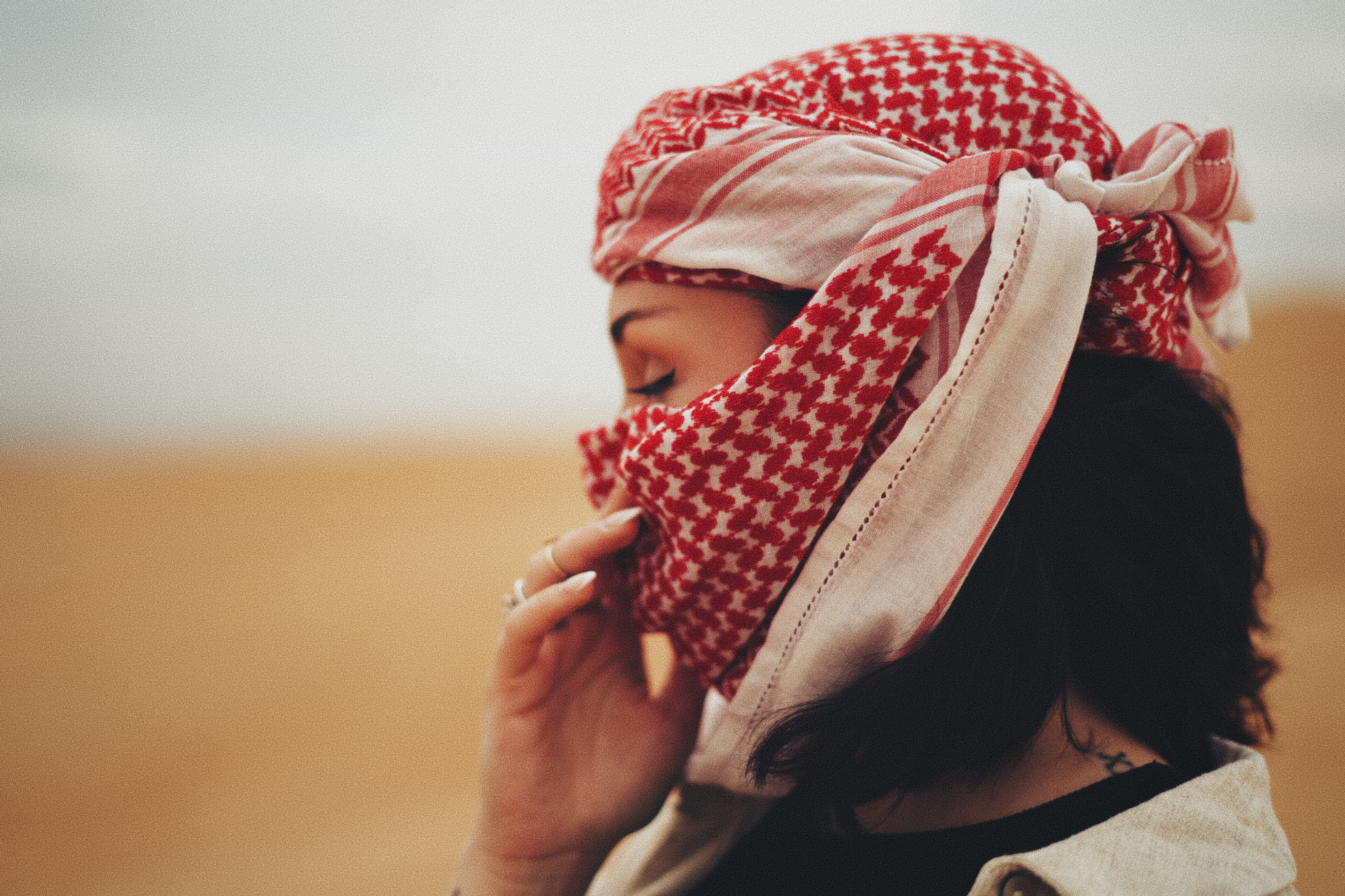 Person wearing a red and white patterned keffiyeh covering their head and lower face, standing in a desert landscape.