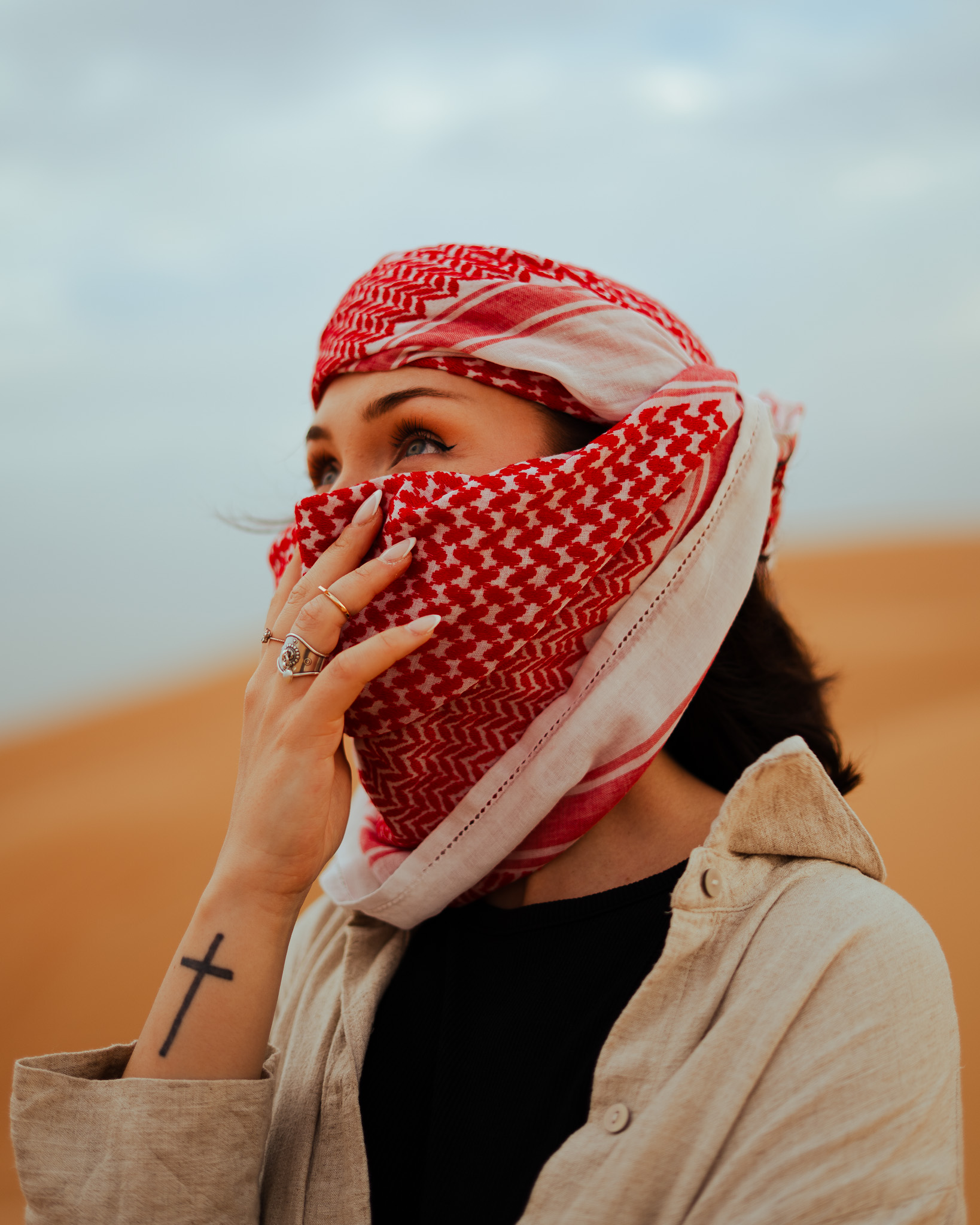Woman wearing a red and white patterned keffiyeh covering her face, with a cross tattoo on her forearm and rings on her fingers.