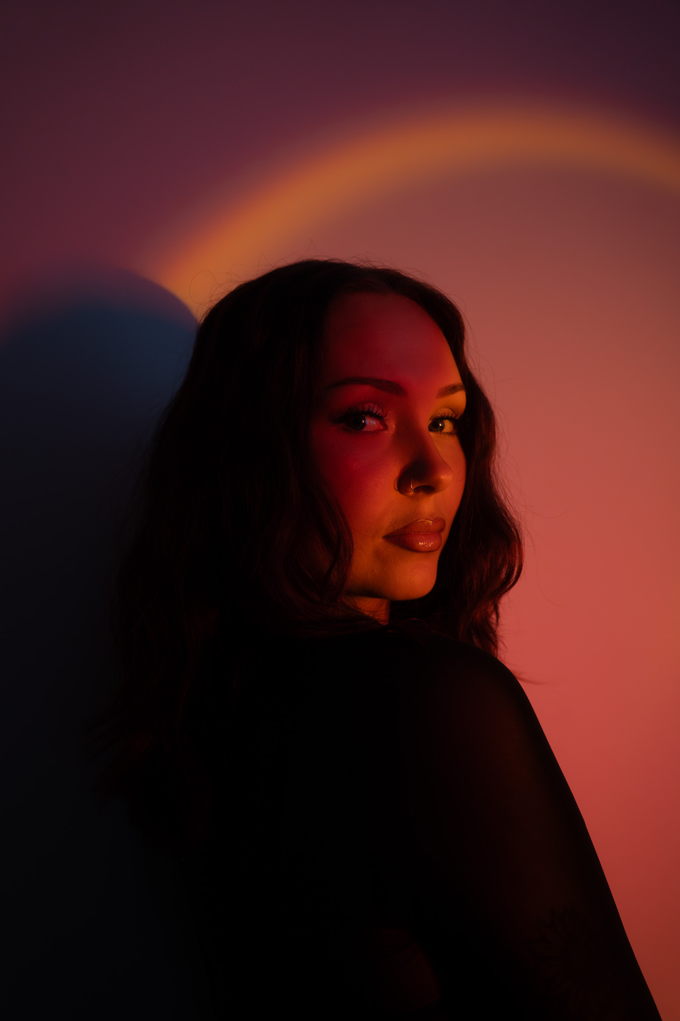 Woman with long dark hair and nose ring looking over her shoulder, illuminated by warm red and orange lighting against a dark background.