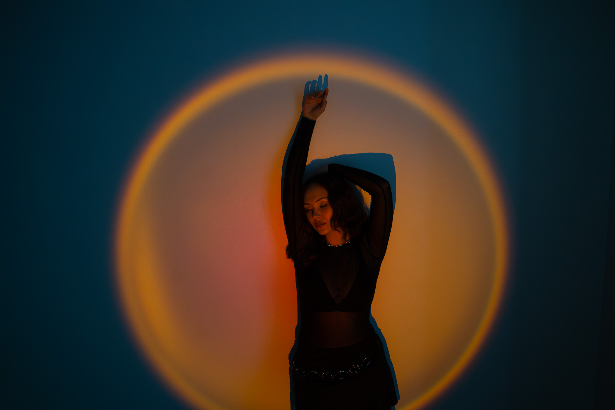 Woman in black dress posing with one arm raised inside a glowing circular light projection on a dark background.