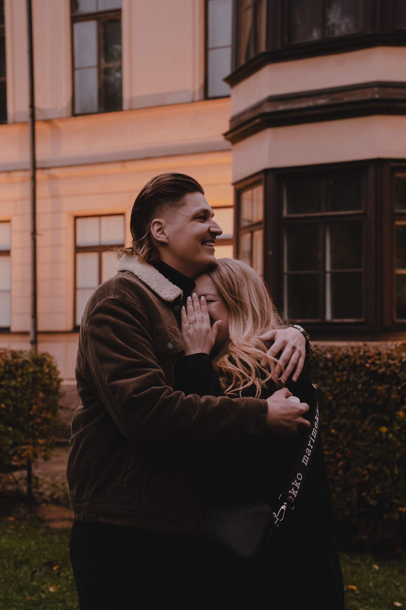 Couple embracing outdoors in front of a building at dusk, with the woman covering part of her face with her hand.