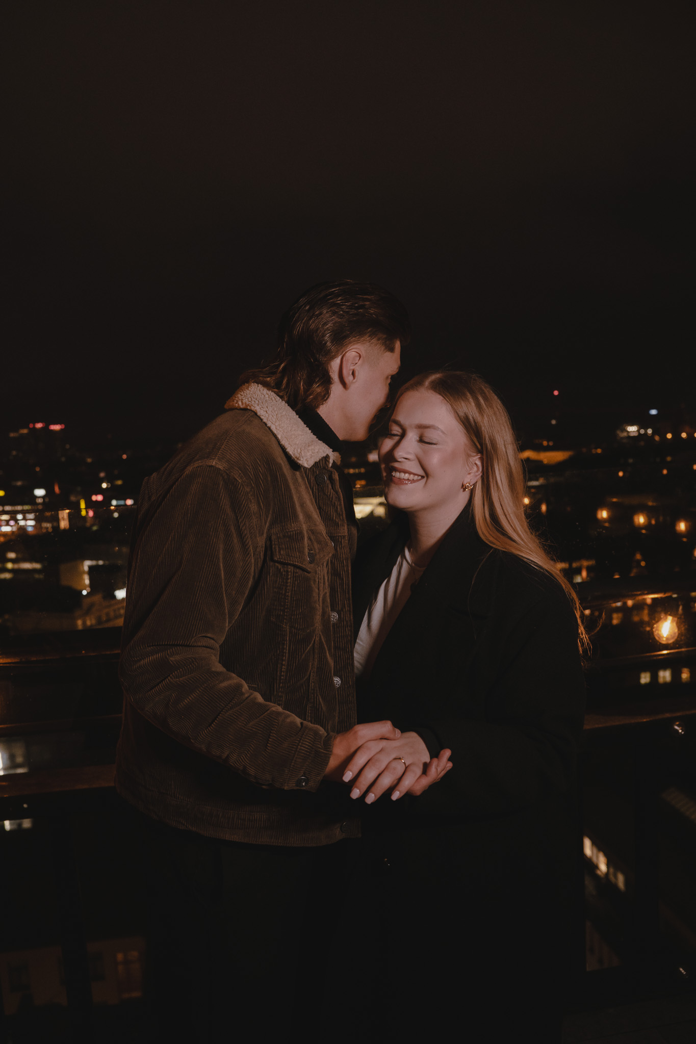 Couple holding hands closely on a rooftop at night with city lights in the background.