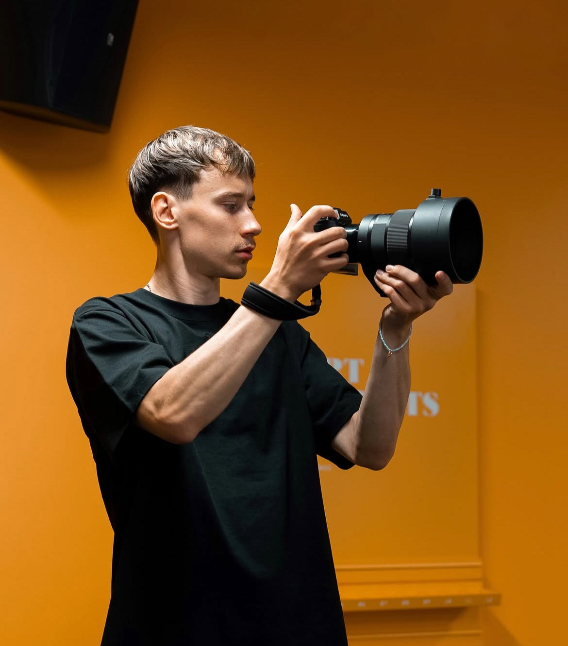 Young man with short hair holding a camera with a large telephoto lens, standing against an orange wall.