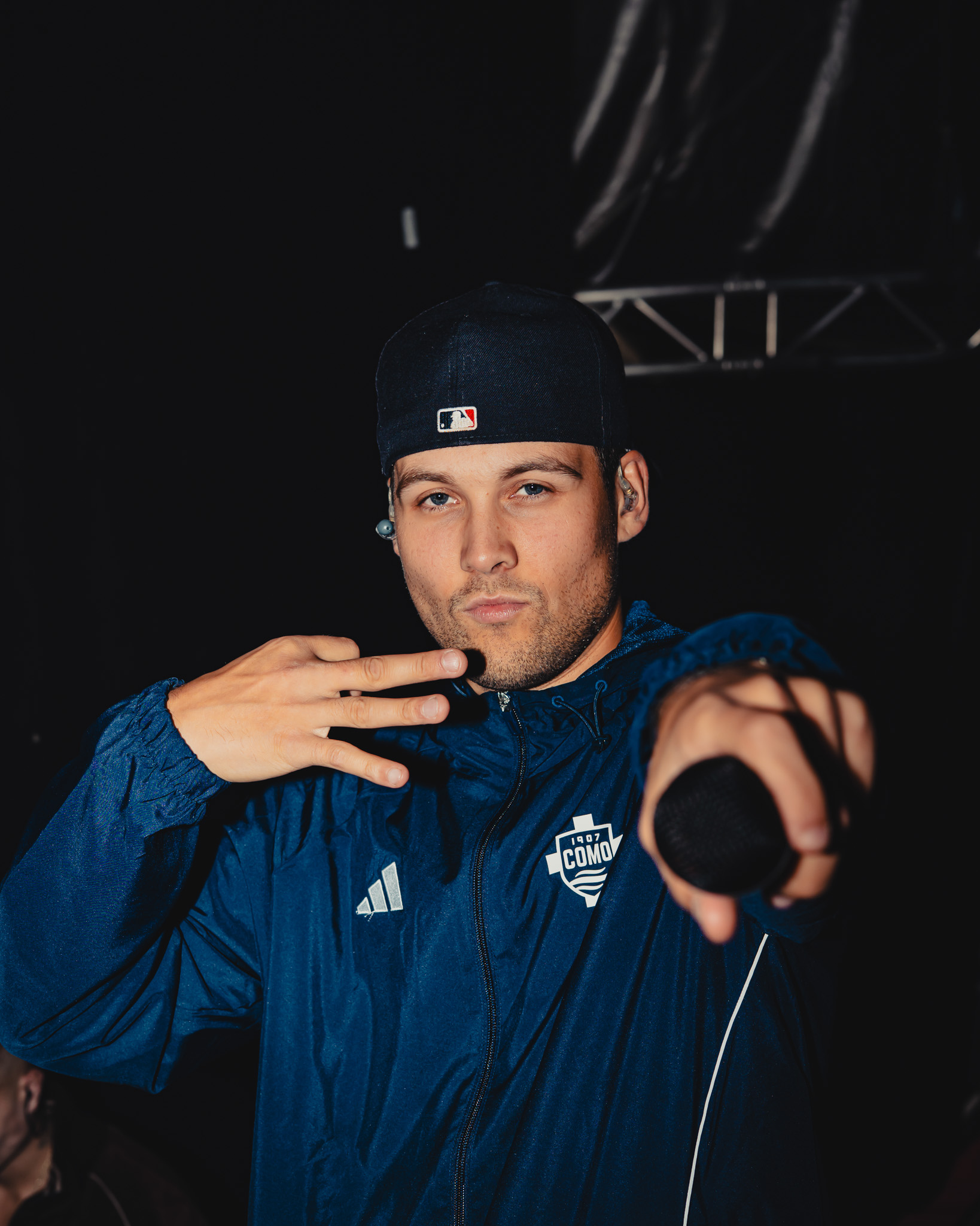 Man in navy blue jacket and backward baseball cap pointing a microphone towards the camera against a dark background.