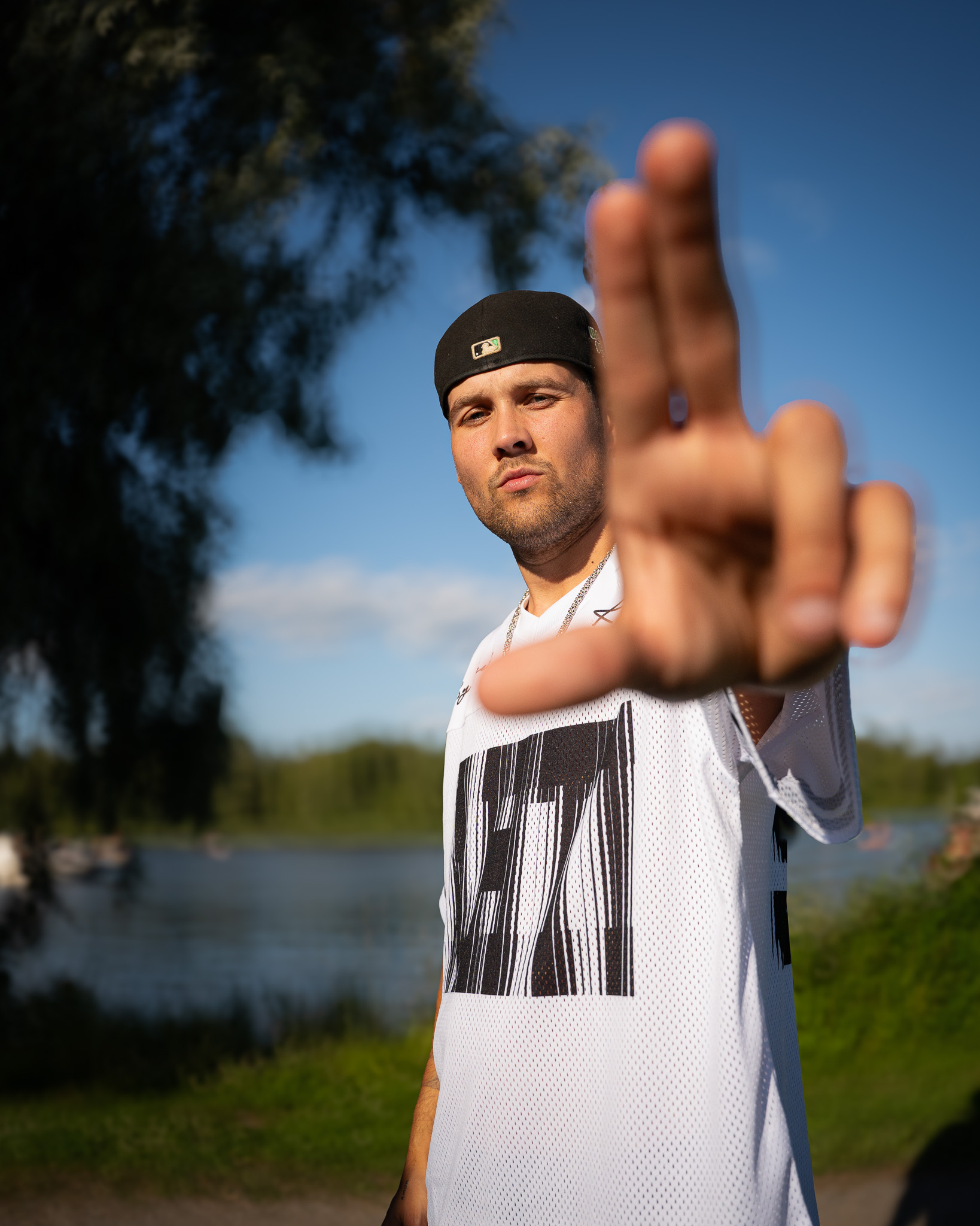 Man wearing a black baseball cap and white sports jersey making an L shape with his fingers outdoors in front of a lake and trees.