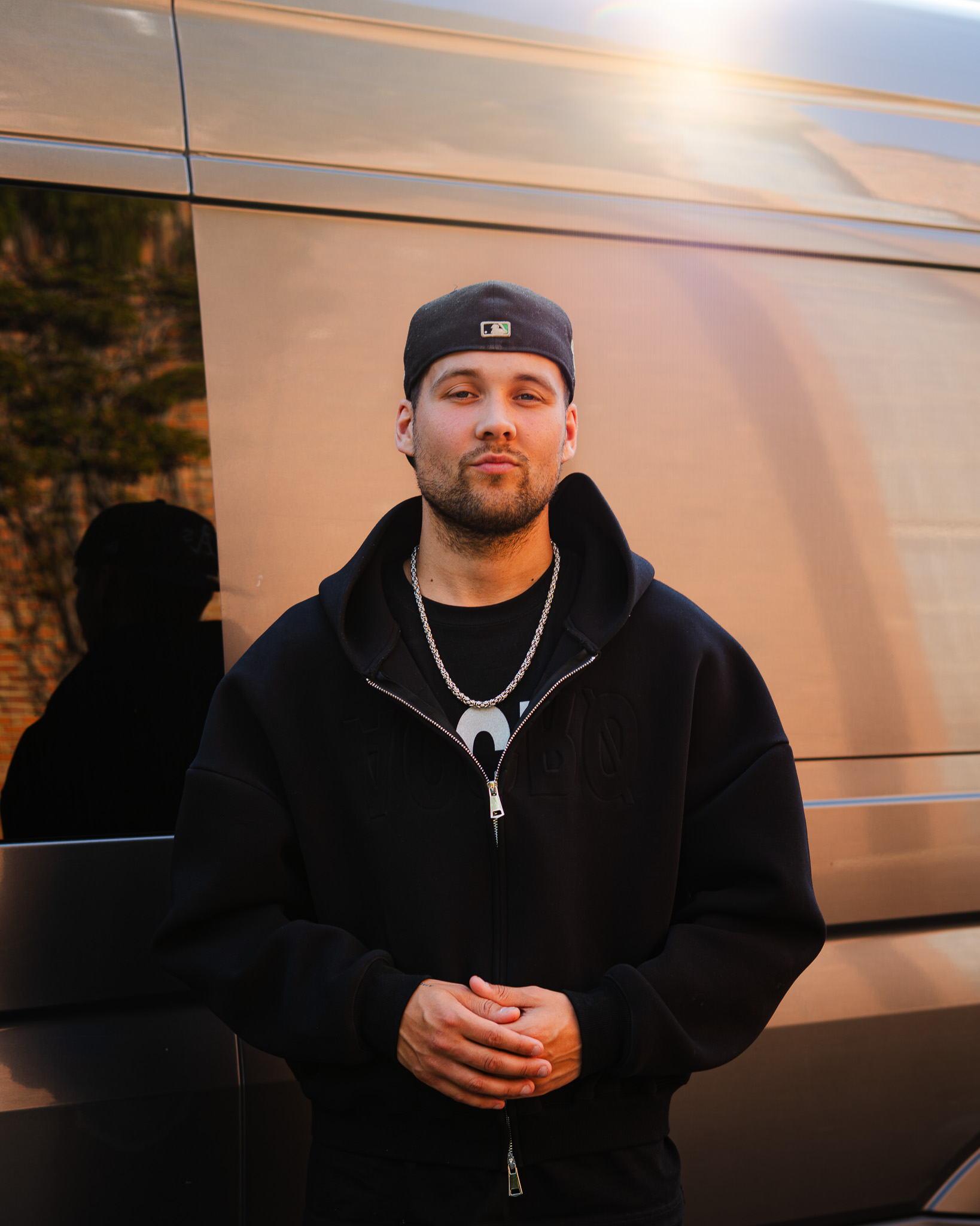 Man wearing a black cap backwards, black hoodie, and silver chain standing in front of a metallic van.