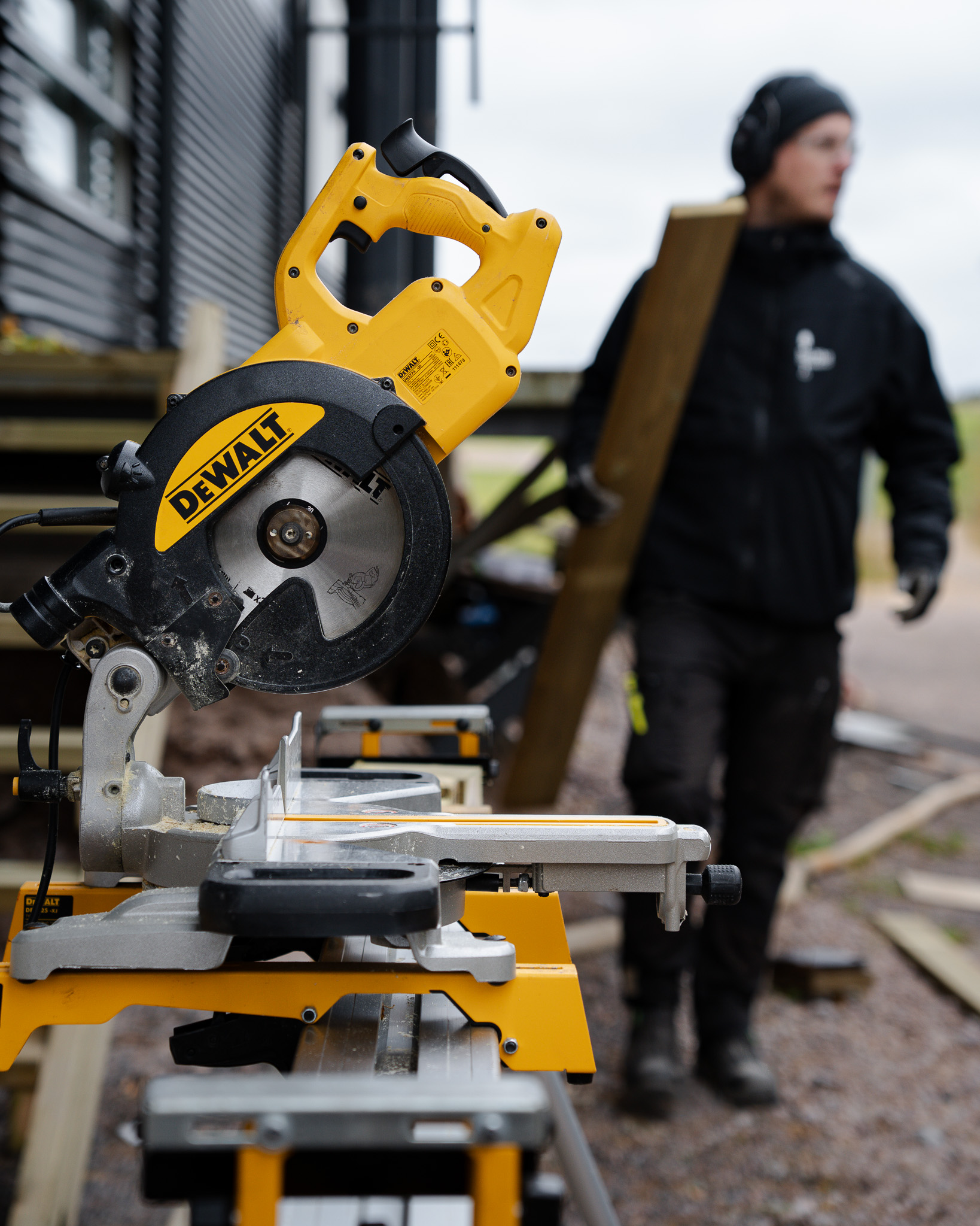 Close-up of a yellow and black DEWALT miter saw on a stand with a blurred person carrying a wooden plank in the background.