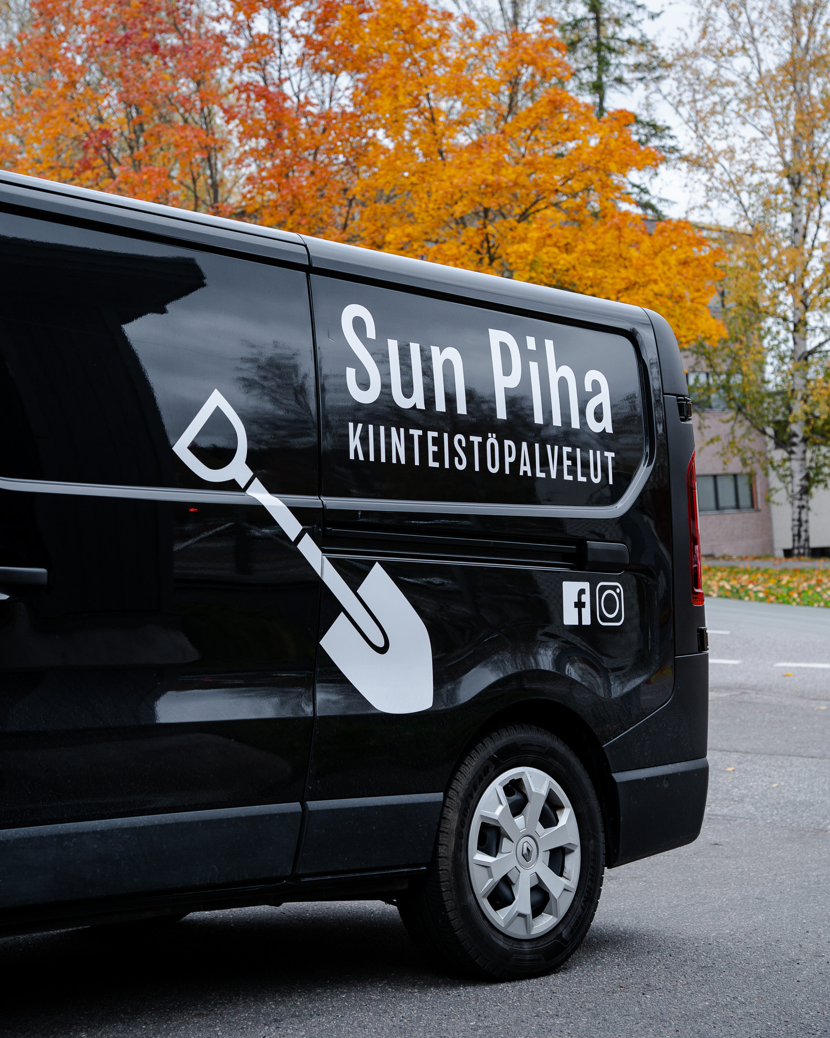 Black service van with white text 'Sun Piha Kiinteistöpalvelut' and a shovel graphic parked on a street with autumn trees in the background.