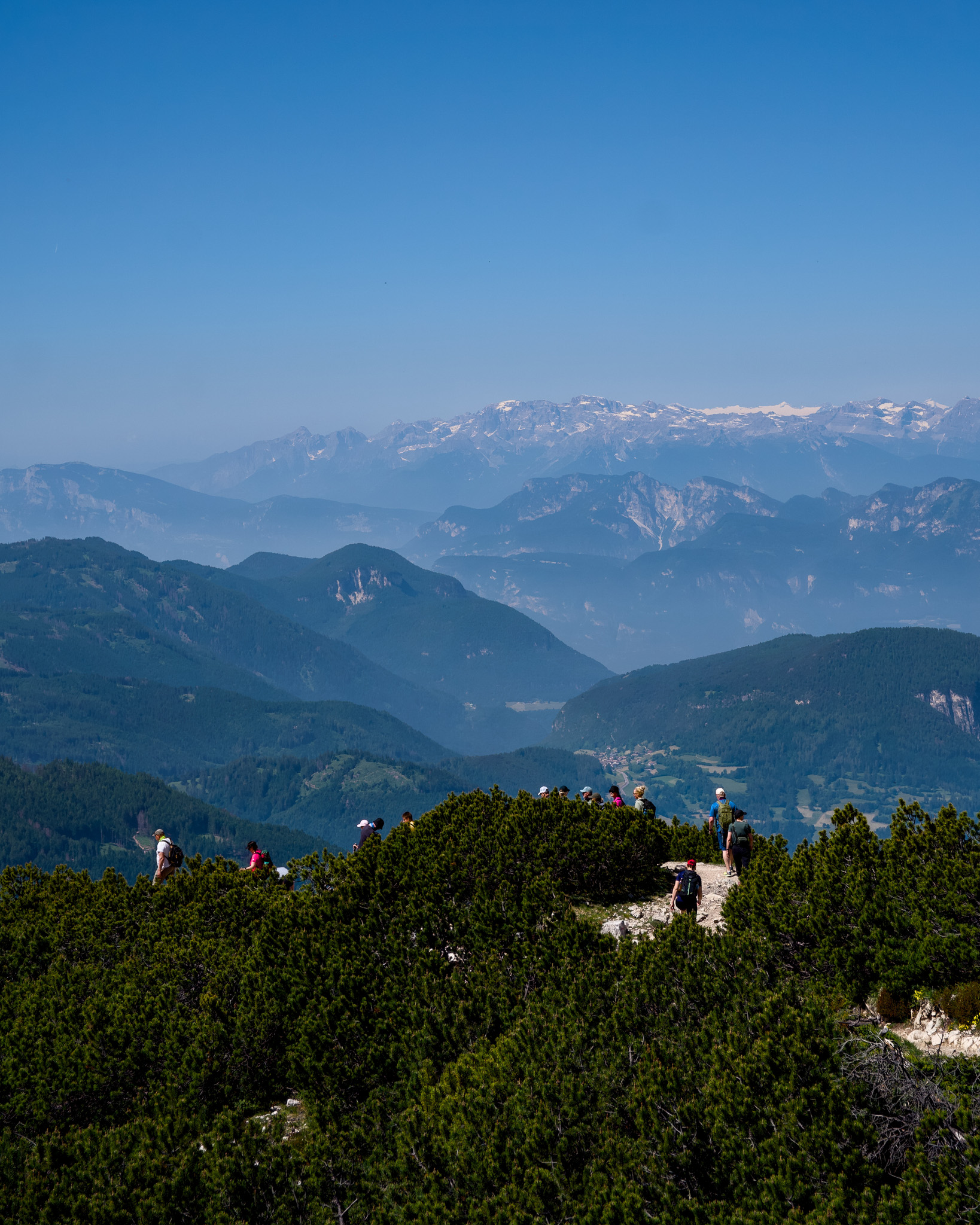 Group of hikers walking along a mountain ridge with layers of forested hills and distant snow-capped peaks under a clear blue sky.