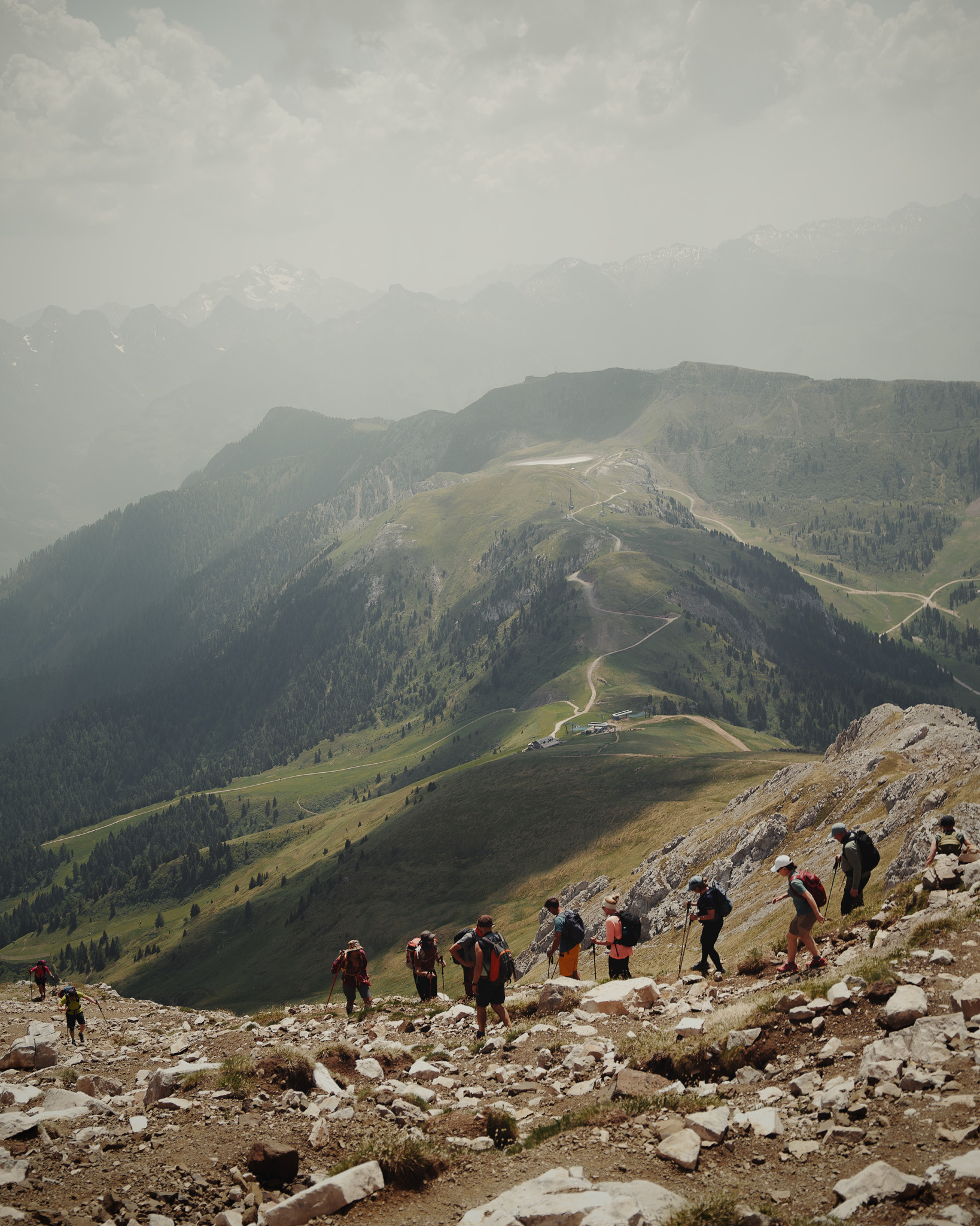 Group of hikers climbing a rocky mountain trail with expansive green valleys and misty mountain peaks in the background.