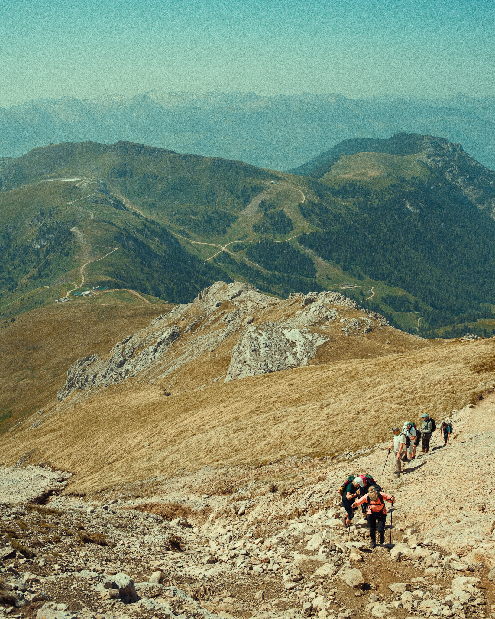 Group of hikers ascending a rocky mountain trail with rolling hills and distant mountain ranges in the background under clear sky.