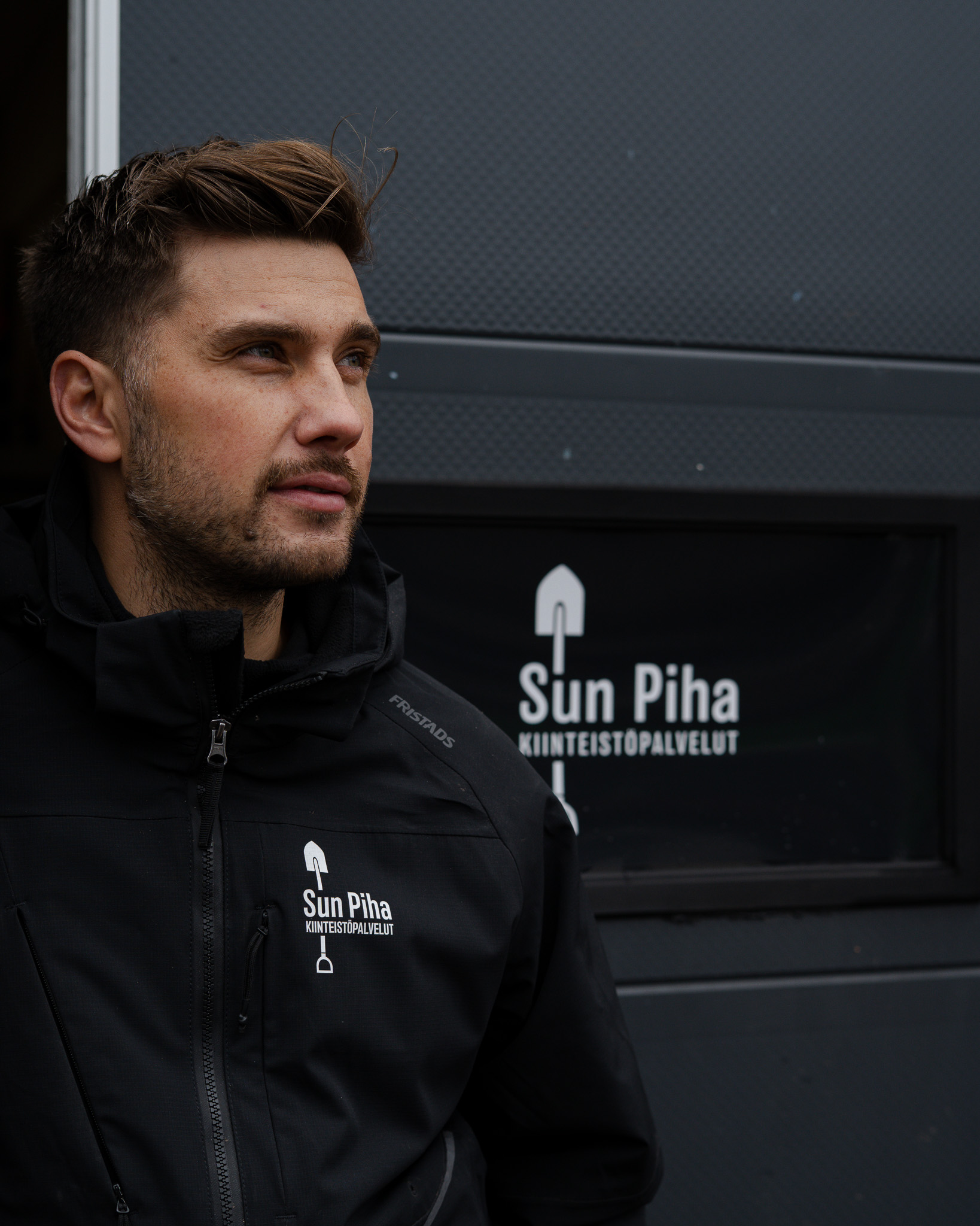 Man with short hair and stubble wearing a black jacket with Sun Piha Kiinteistöpalvelut logo, standing in front of a door with matching signage.