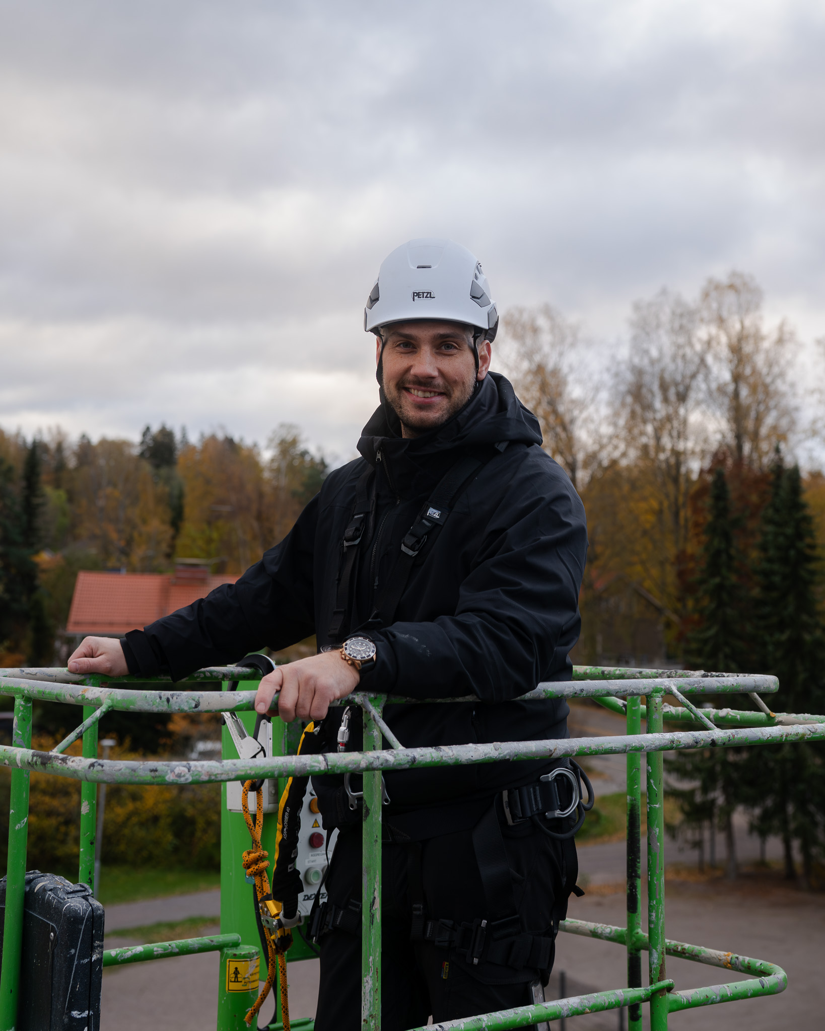 Man wearing a white safety helmet and black jacket standing in a green metal safety cage outdoors.