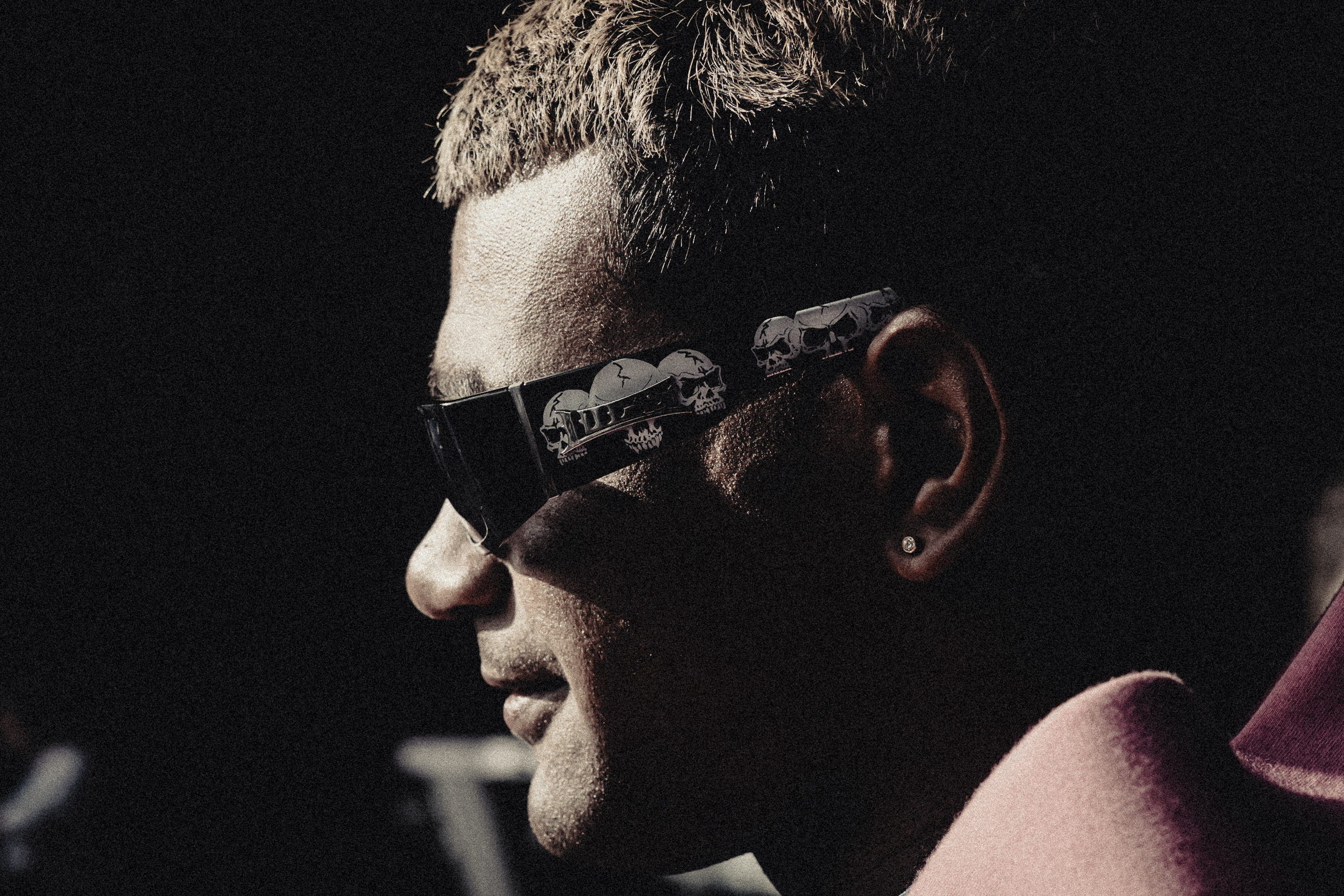 Profile of a man wearing sunglasses decorated with skull designs and a pink shirt, against a dark background.