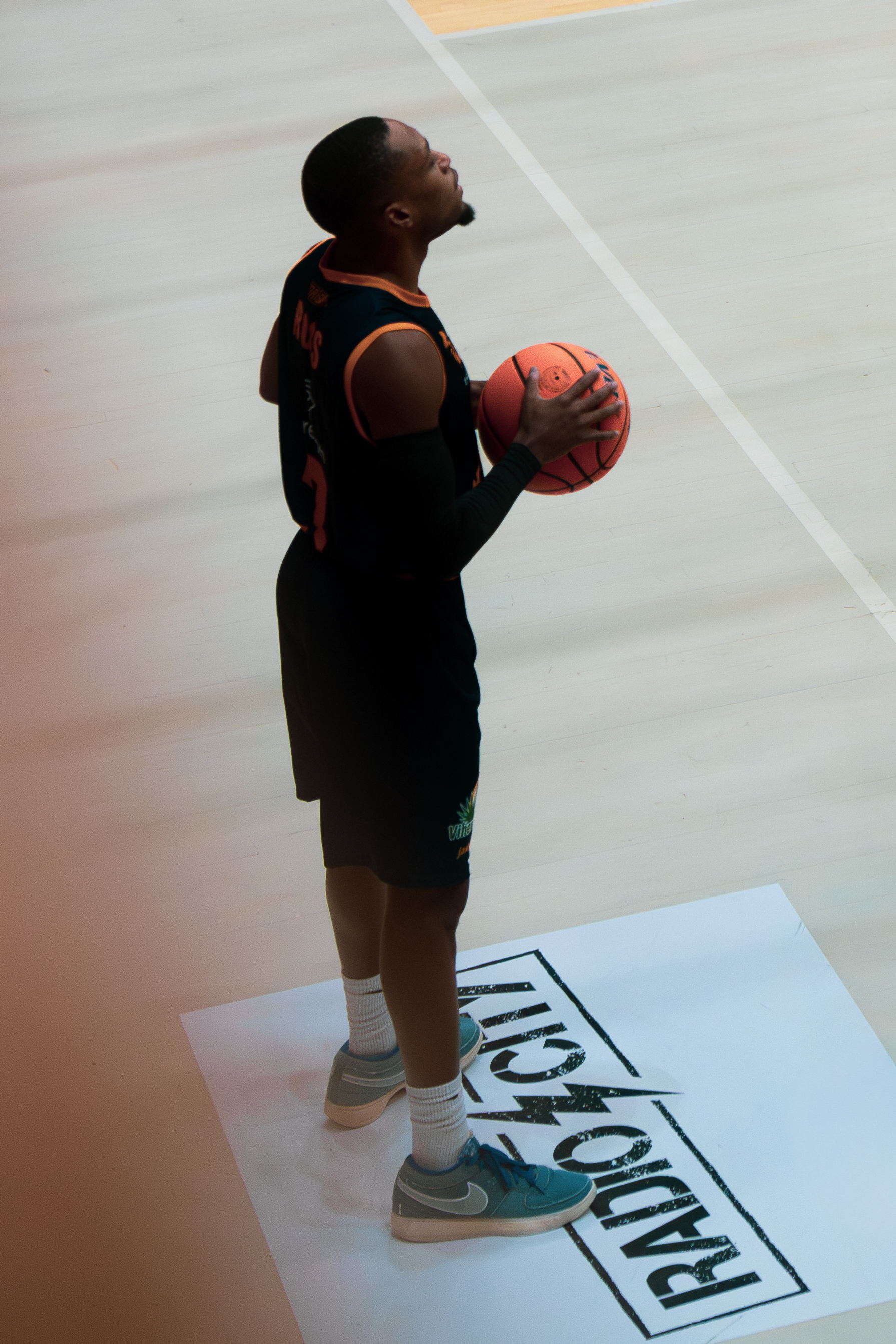 Basketball player in black uniform holding a basketball, standing on a court near a 'RADIO SHACK' logo mat.