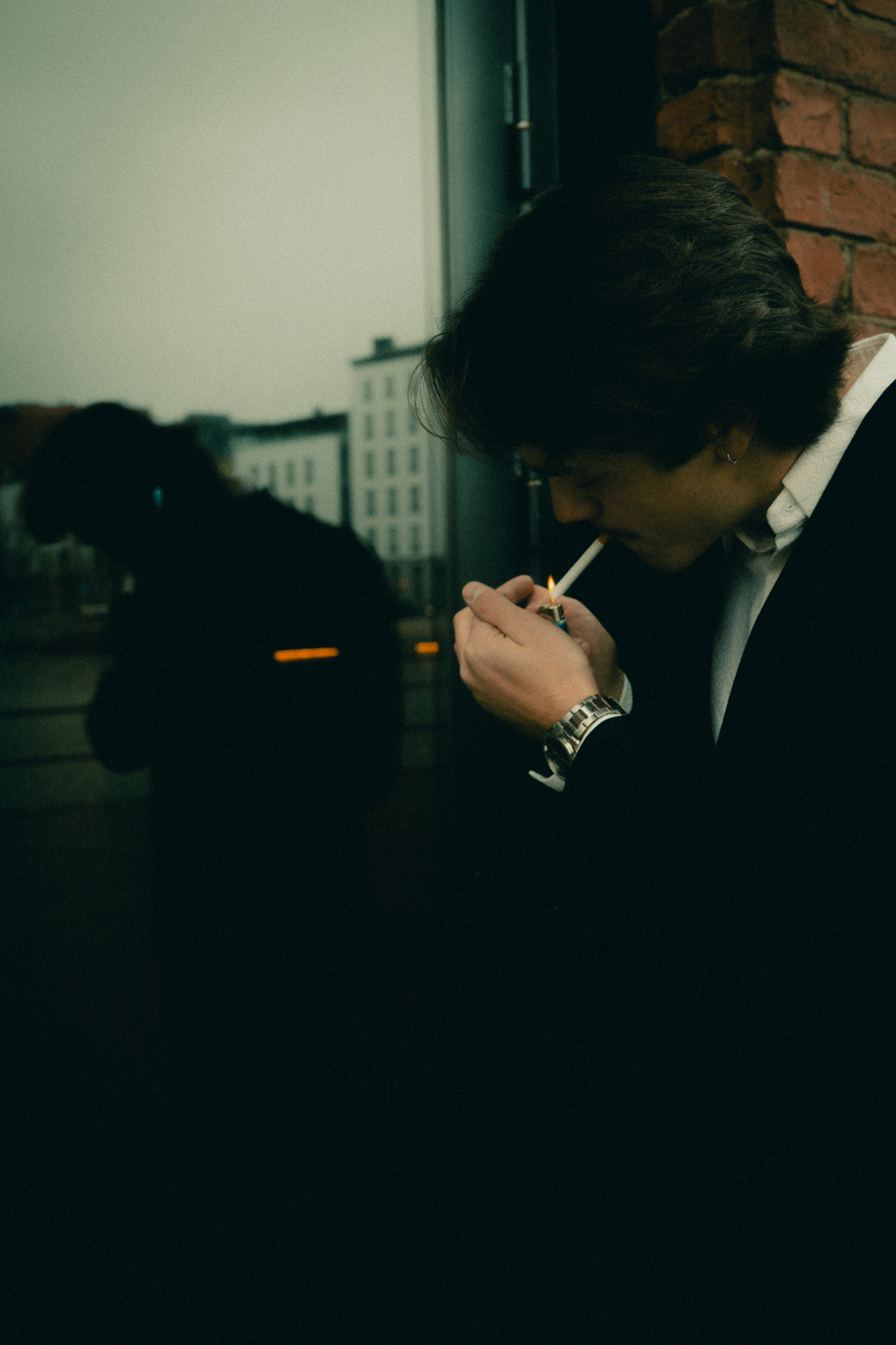 Young man lighting a cigarette outdoors next to a brick wall with a city building reflected in a glass surface.