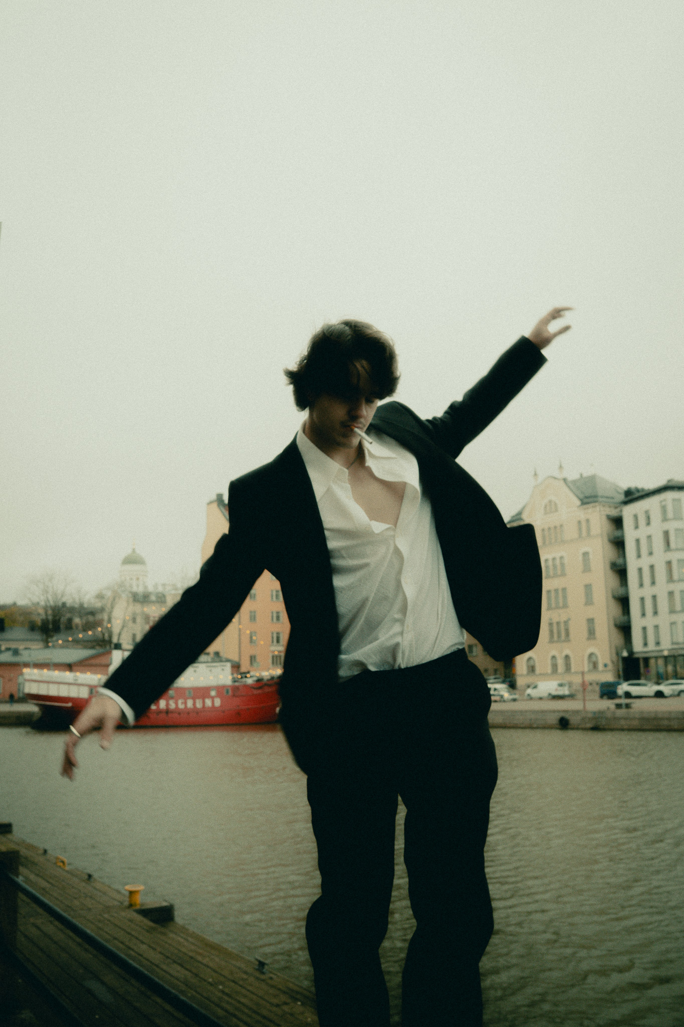 Young man in a black suit with an unbuttoned white shirt poses near a waterfront with buildings and a red boat in the background.