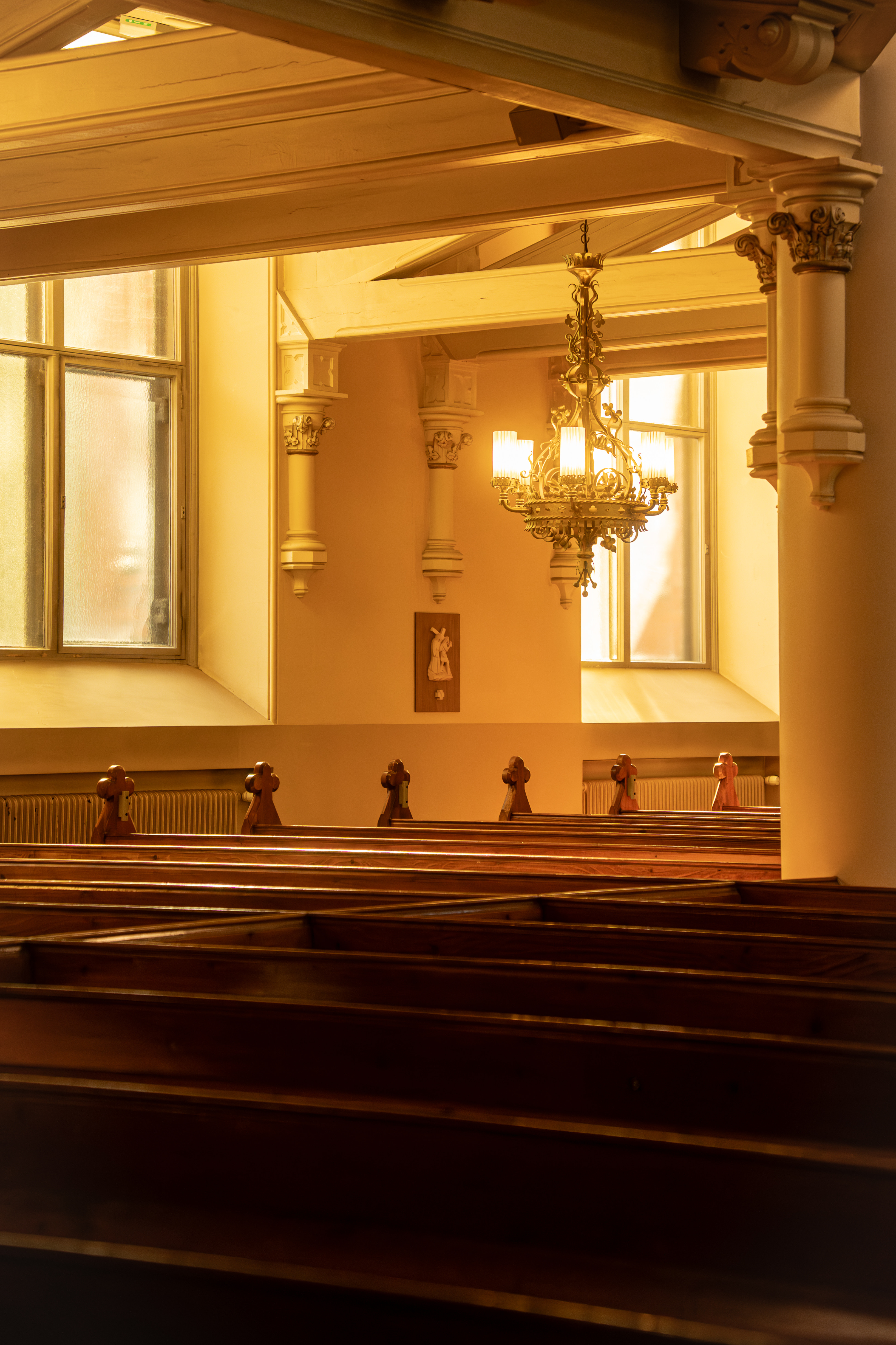 Interior view of a warmly lit church with wooden pews, decorative columns, arched beams, and a hanging chandelier between two tall windows.
