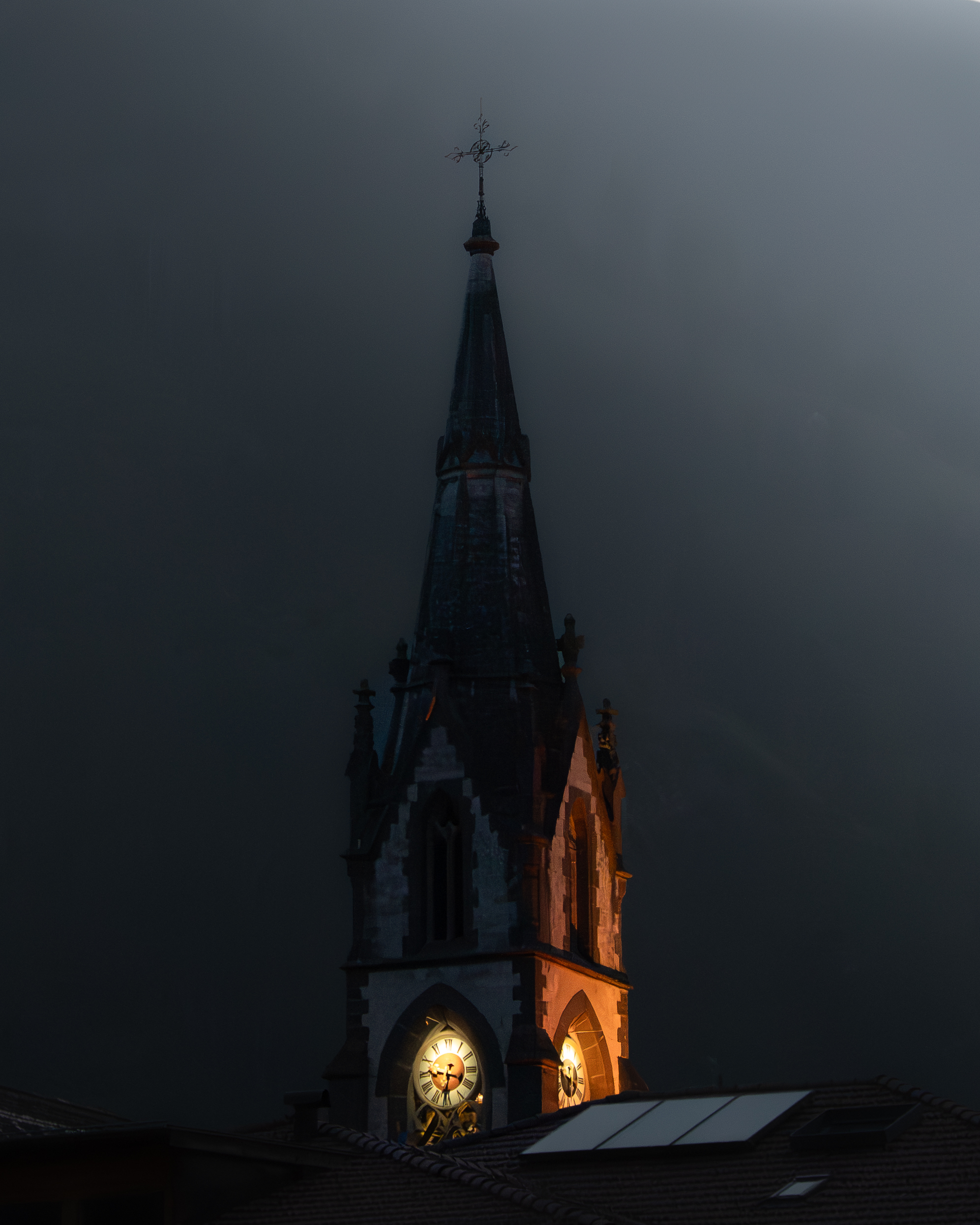 Illuminated clock tower with a pointed steeple and cross at night against a dark sky.