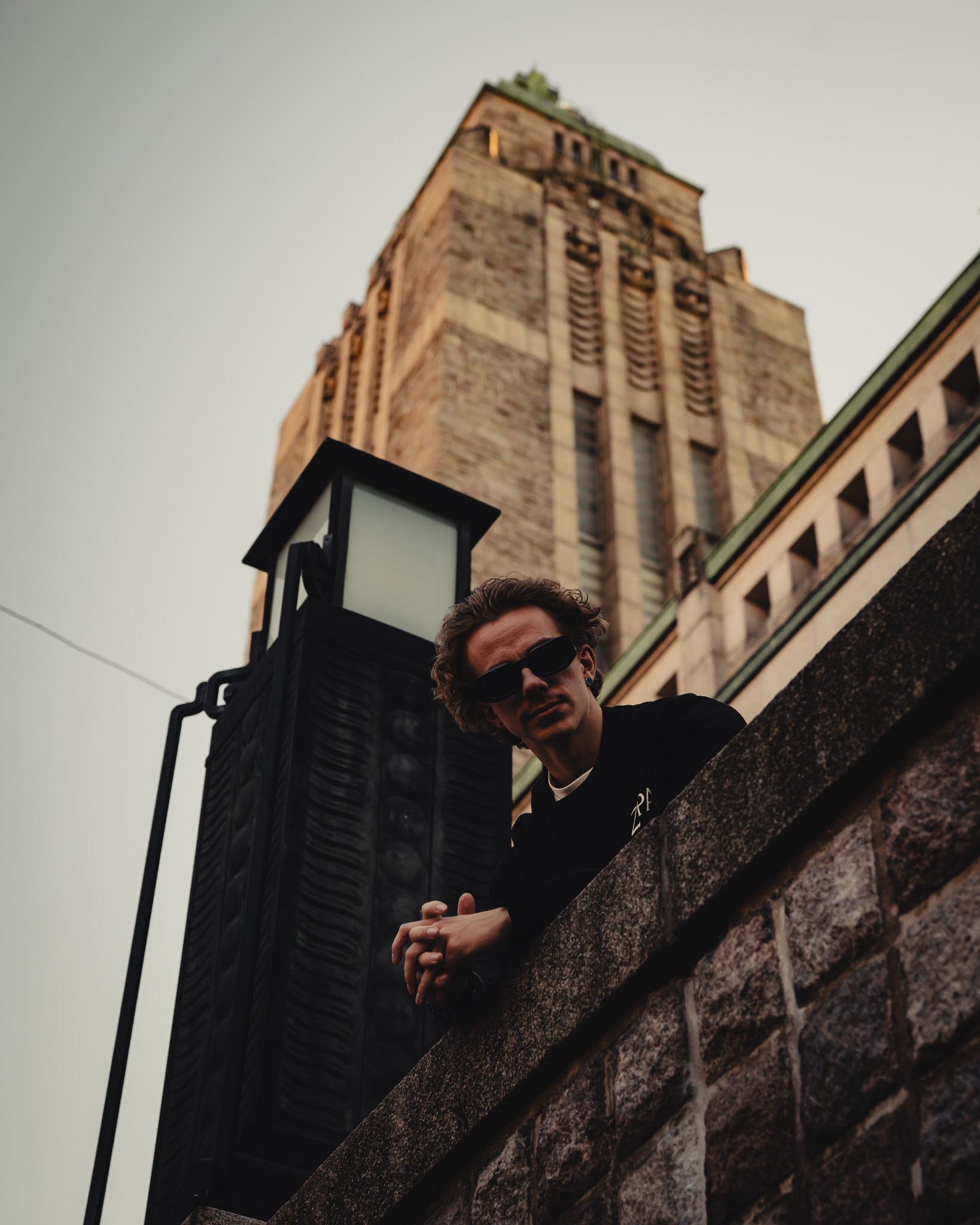 Man wearing sunglasses leaning on a stone ledge with a tall historic building and a black lamp post in the background.