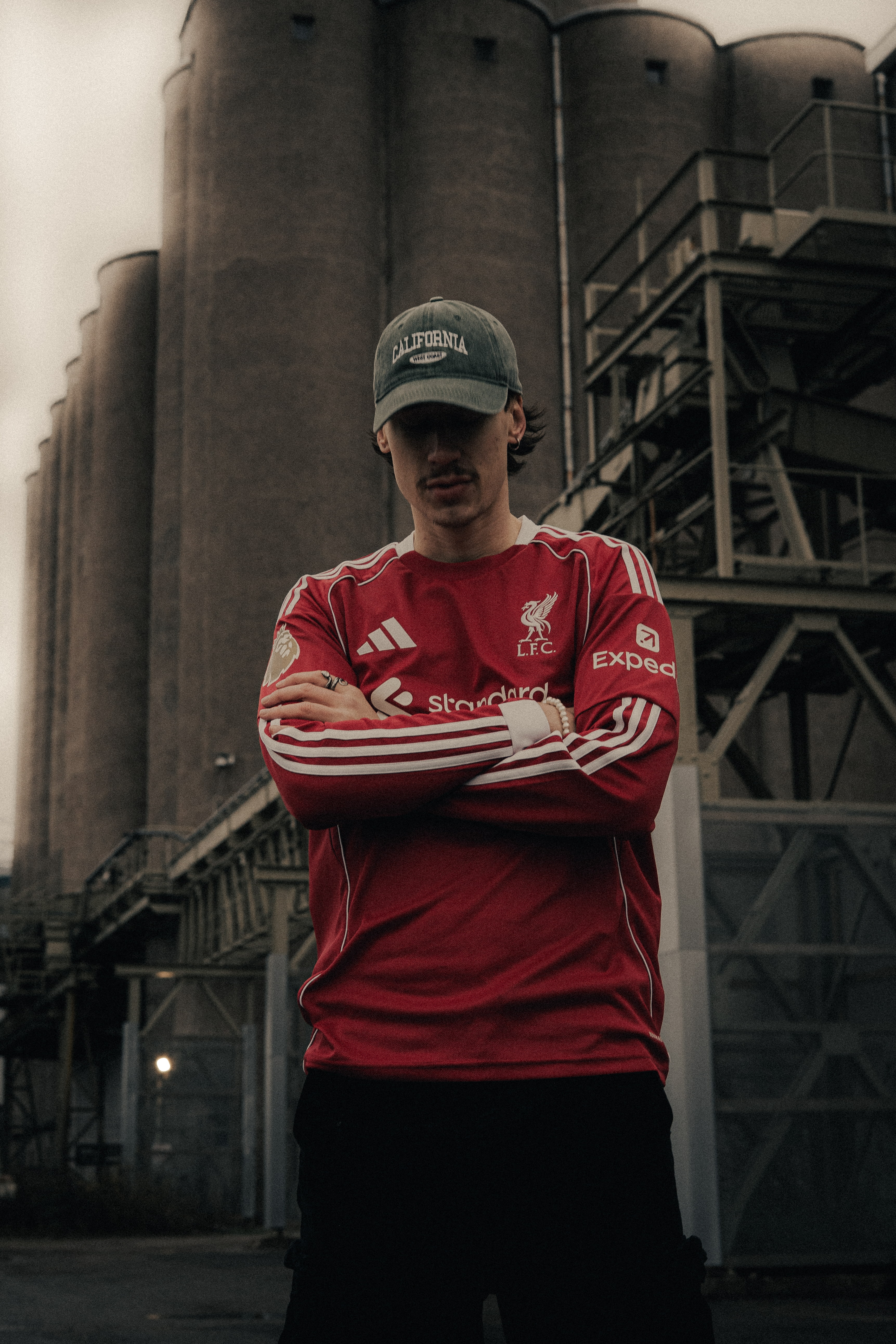 Young man wearing a red Liverpool FC jersey and a green California cap standing with arms crossed in front of industrial silos.