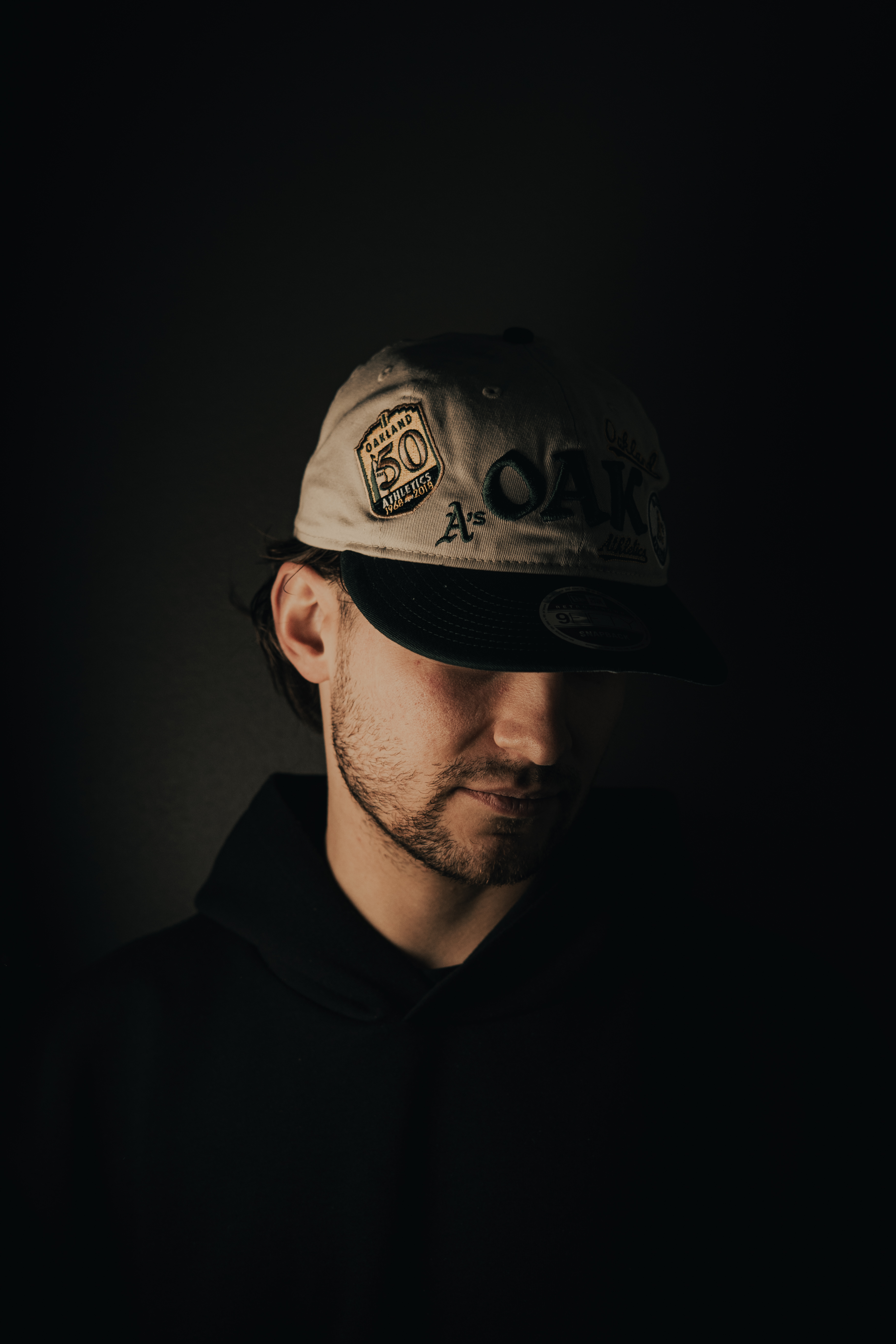 Man wearing an Oakland Athletics baseball cap and a dark hoodie, with face partially shadowed.