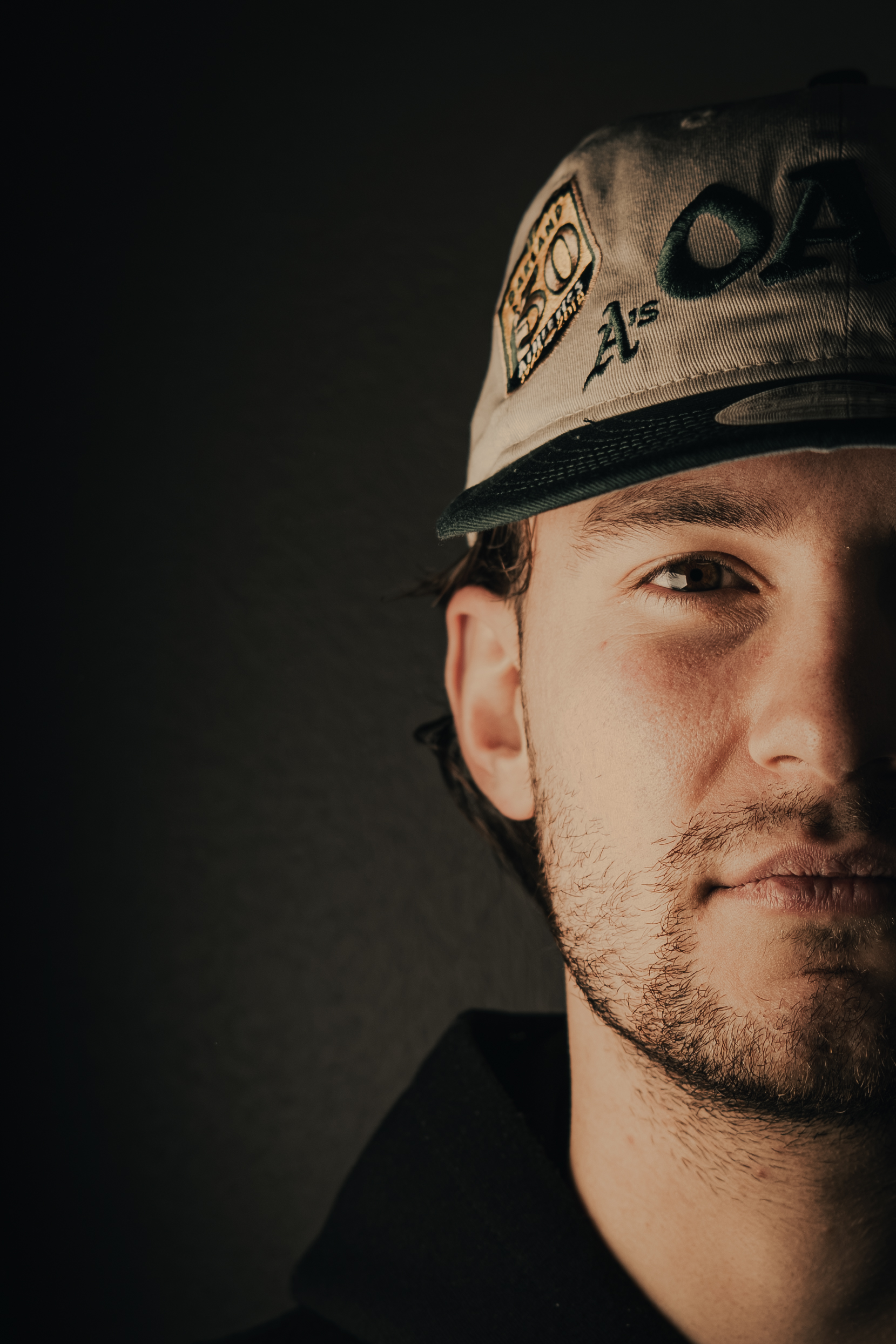 Close-up of a young man with light facial hair wearing an Oakland Athletics baseball cap and a dark hoodie against a dark background.