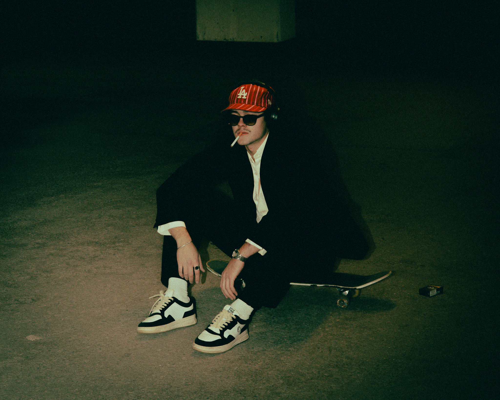 Young man in sunglasses and red LA cap sitting on a skateboard at night with a cigarette in his mouth.