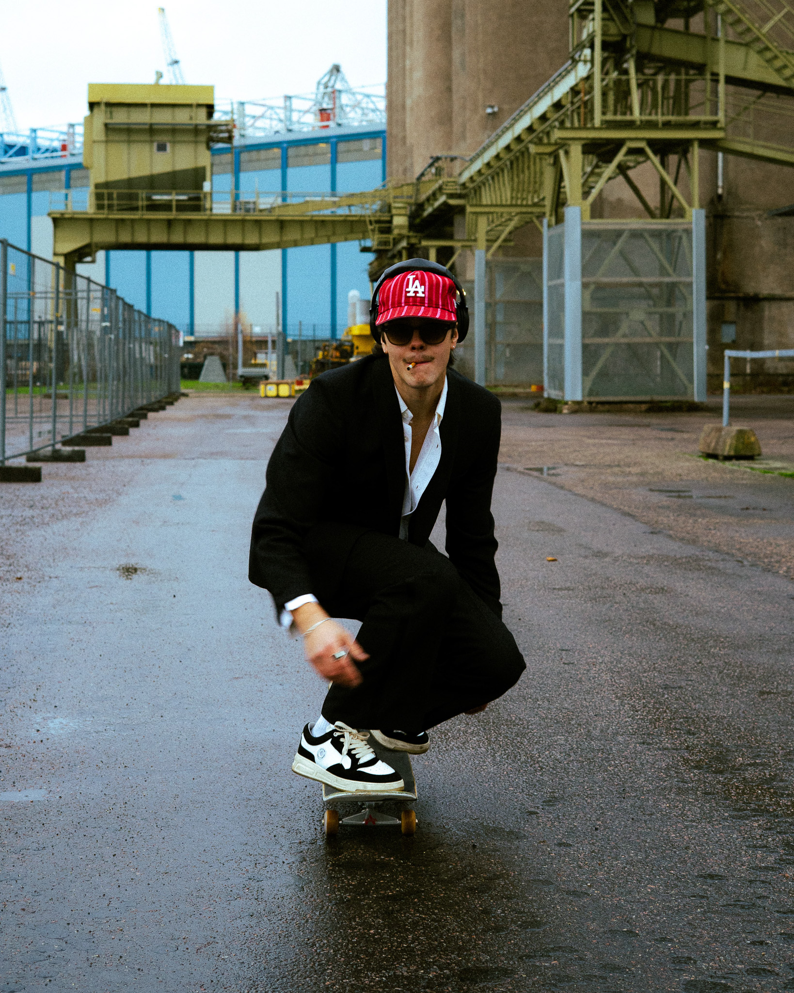 Person wearing a suit, red LA cap, sunglasses, and headphones skateboarding on wet pavement in an industrial area.