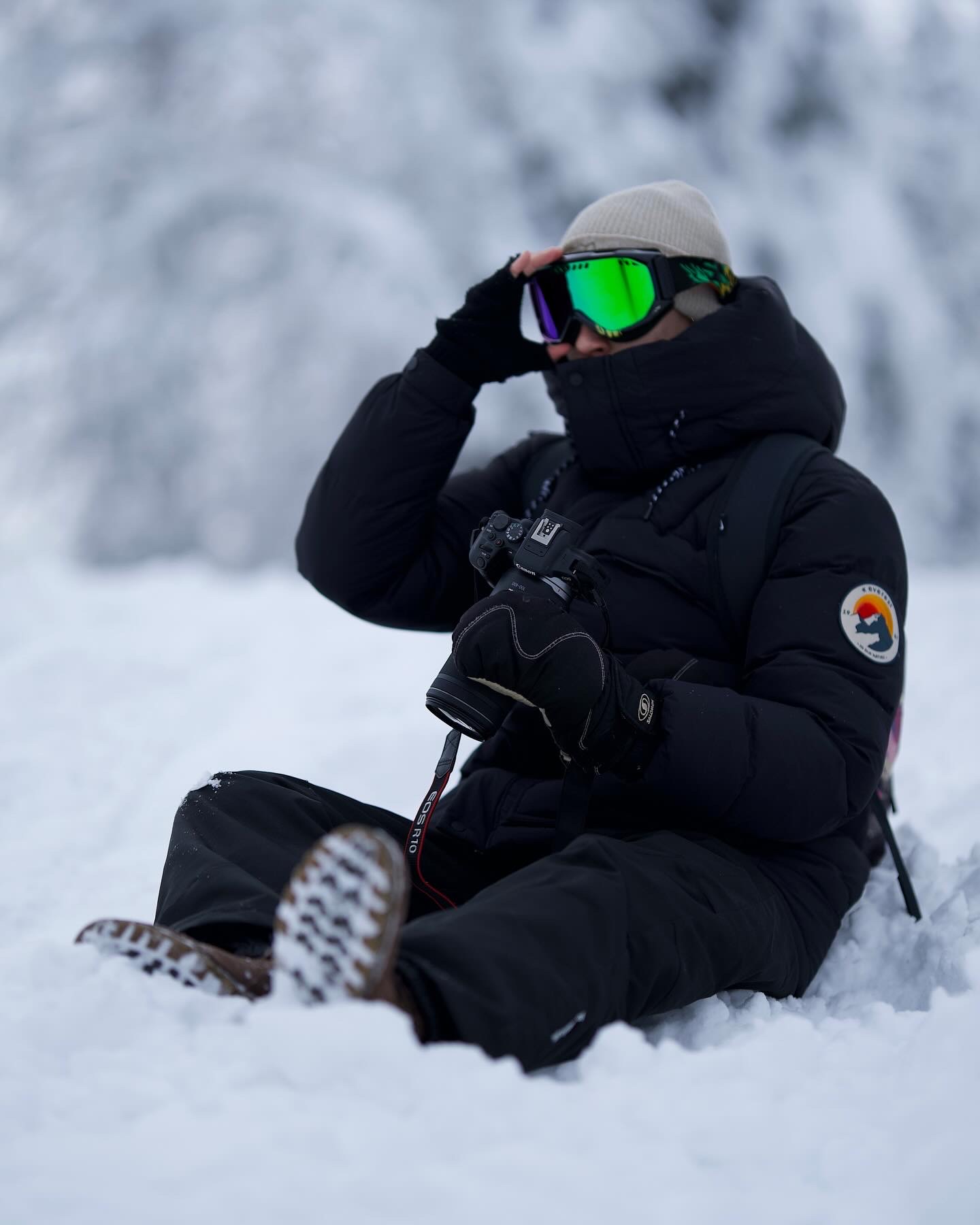 Person dressed in black winter gear with green ski goggles, sitting in the snow holding a camera.
