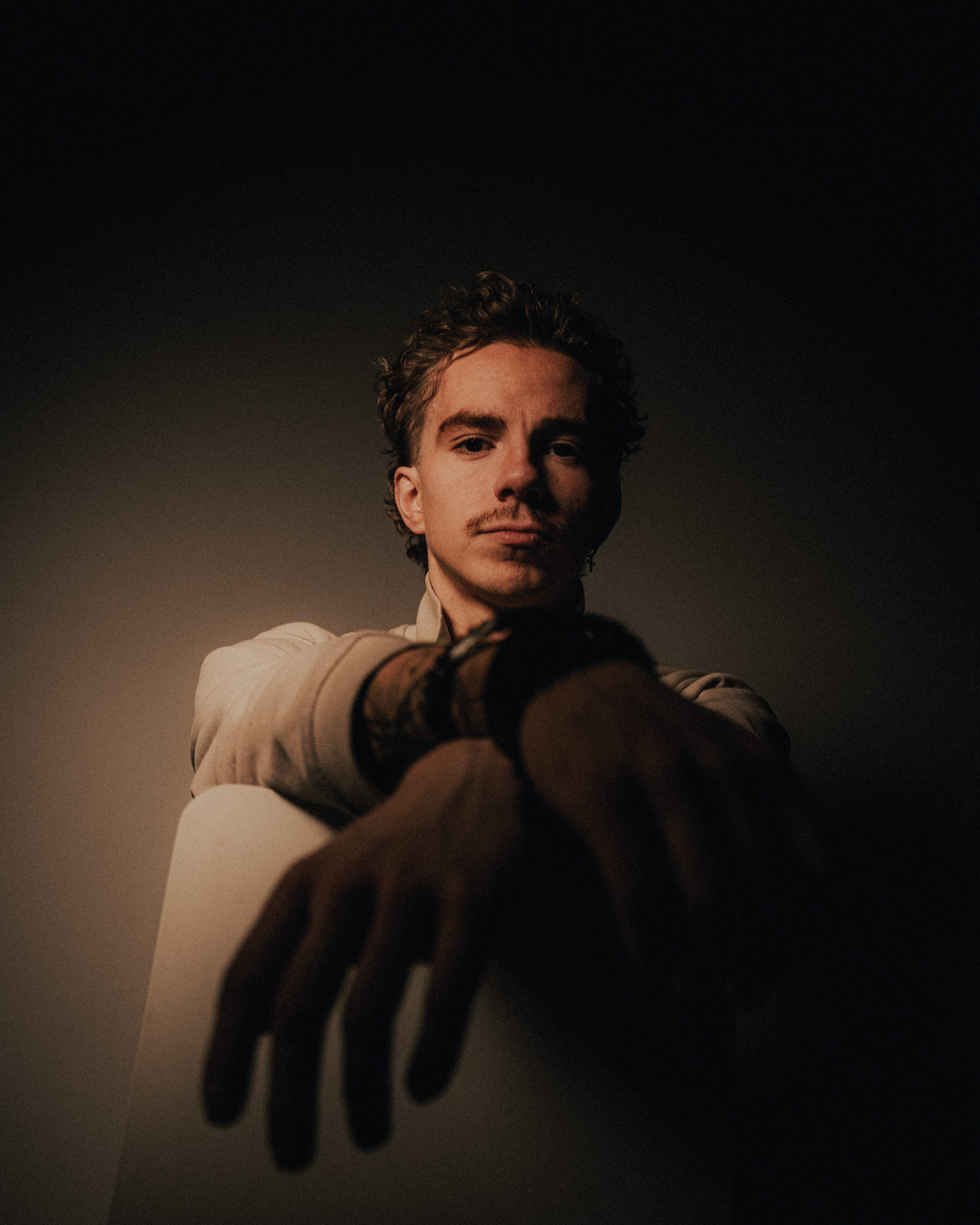 Young man with curly hair and mustache sitting with hands extended toward the camera against a dark background.
