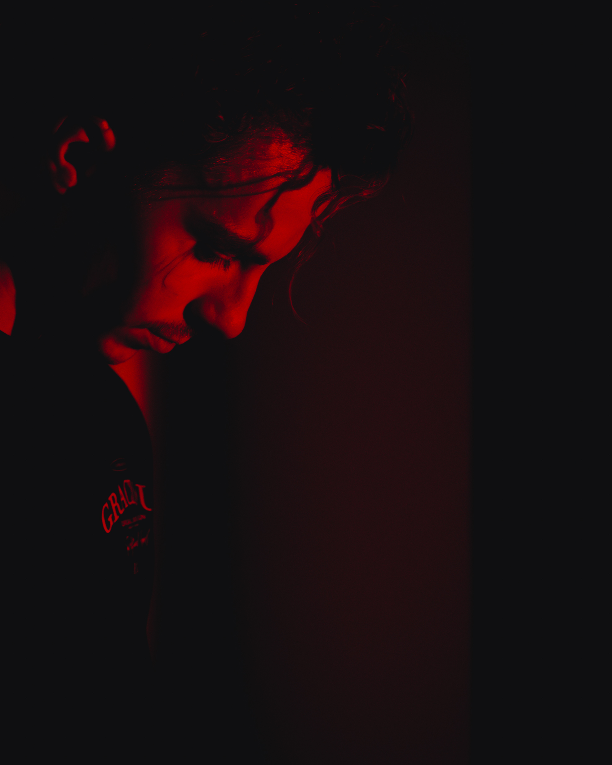 Side profile of a young man with curly hair, illuminated by red light against a dark background.