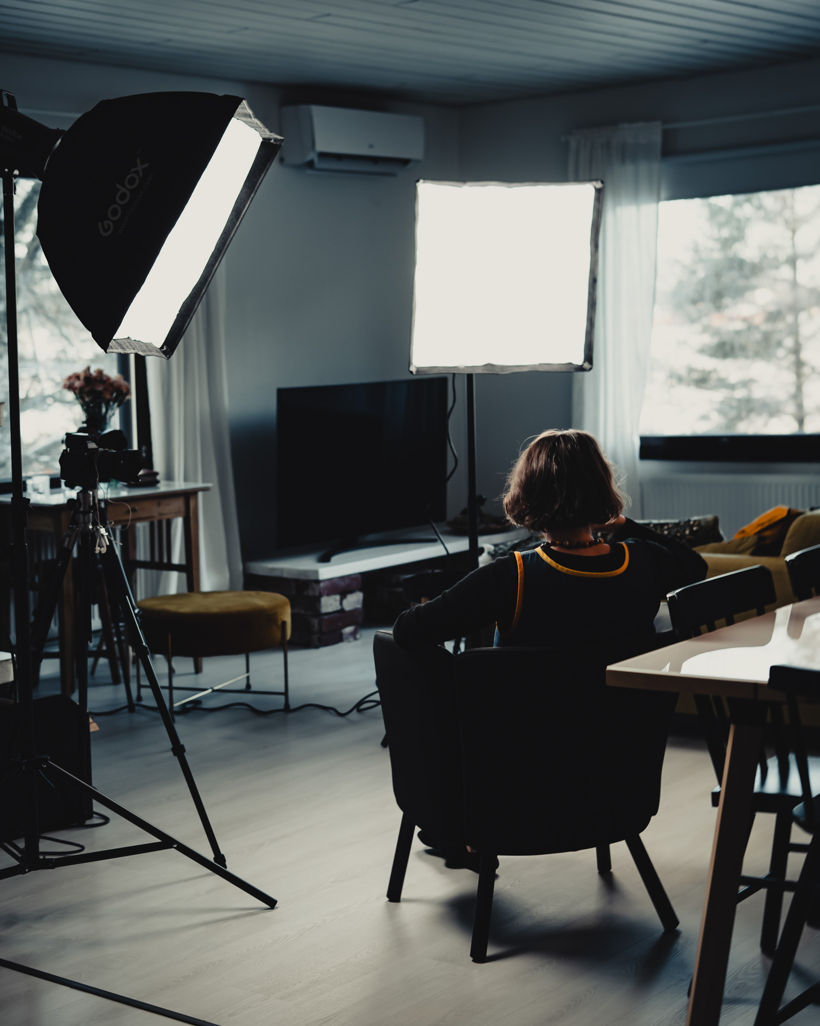Person with short hair sitting on a chair facing a television in a room lit by two large photography softbox lights.