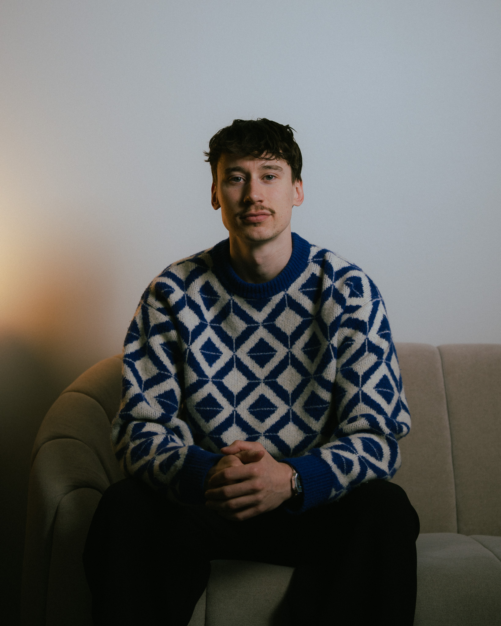 Young man with short curly hair and a mustache sitting on a beige couch wearing a blue and white patterned sweater.