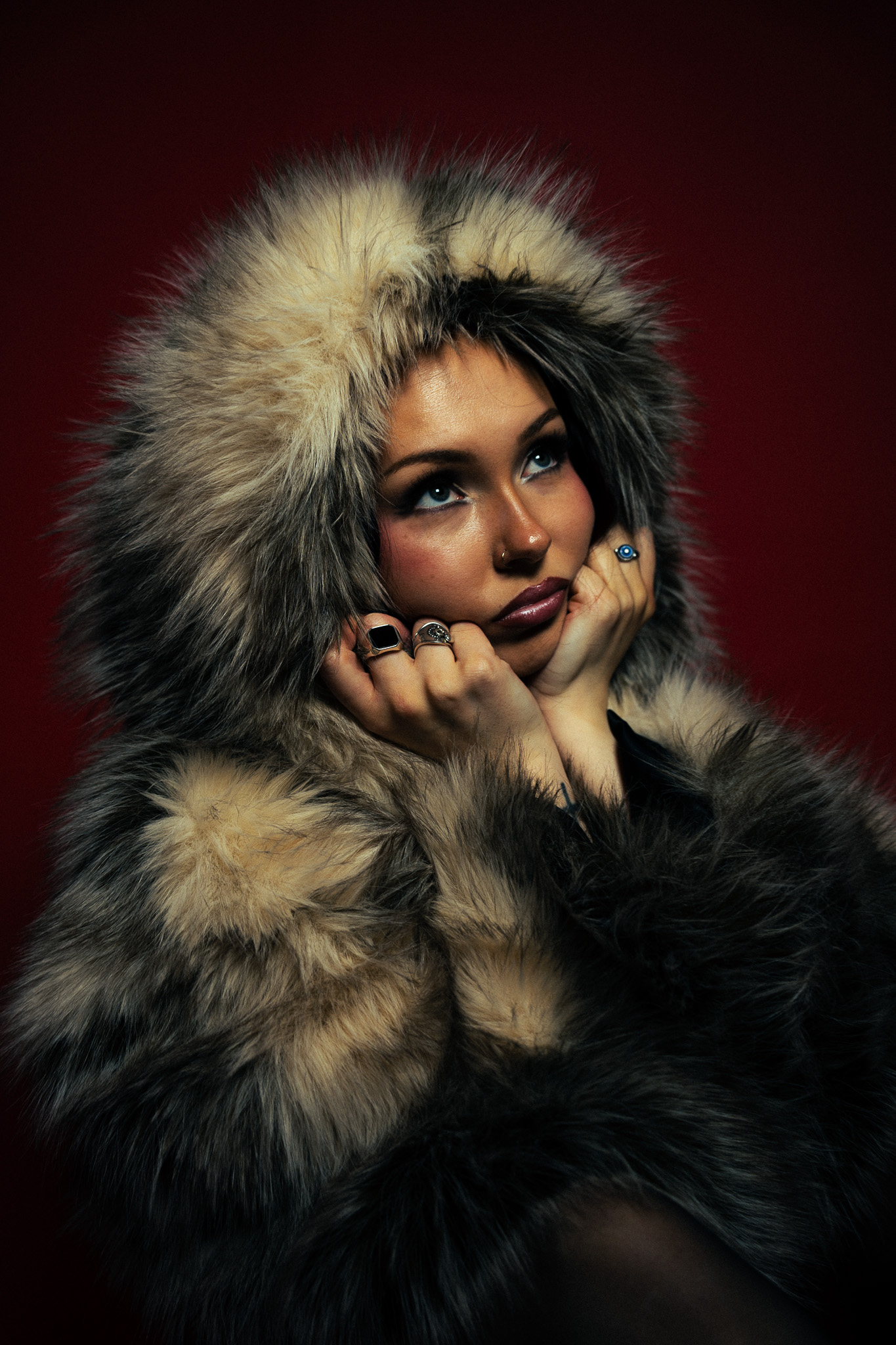 Woman with rings wearing a fluffy fur coat hood and looking thoughtfully upward against a dark red background.
