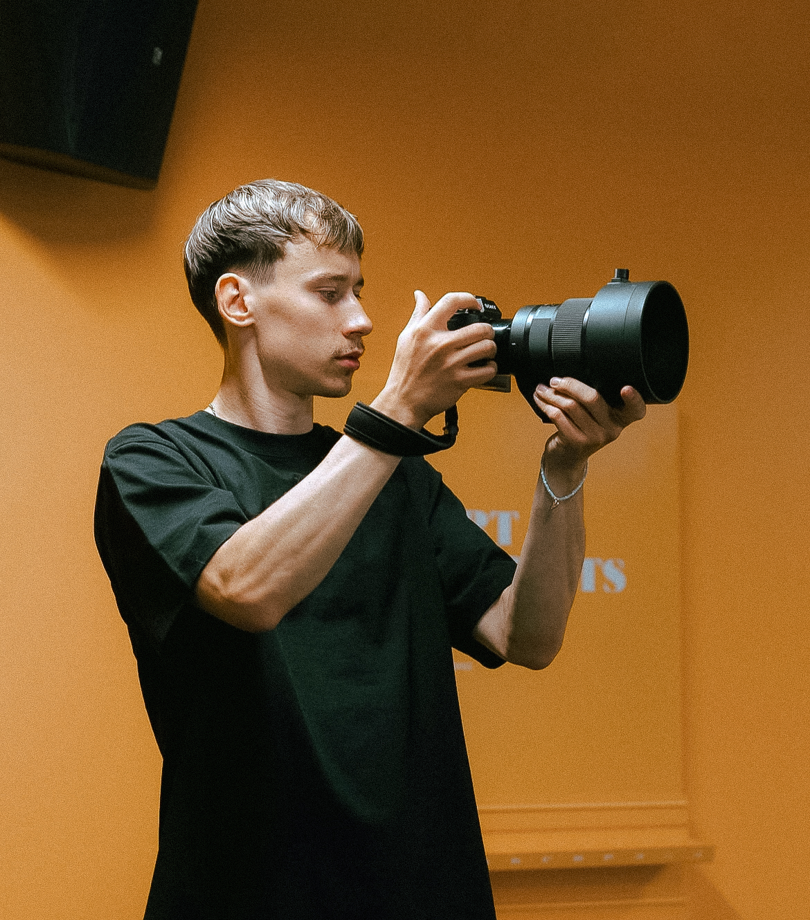 Young man in a black t-shirt holding a camera with a large lens against an orange background.