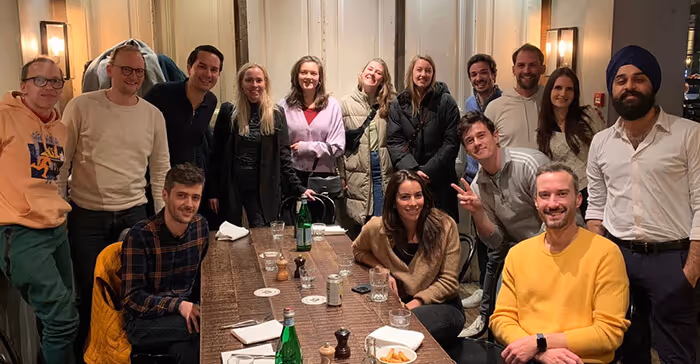 Group of 14 people smiling and posing together around a wooden table in a warmly lit room.