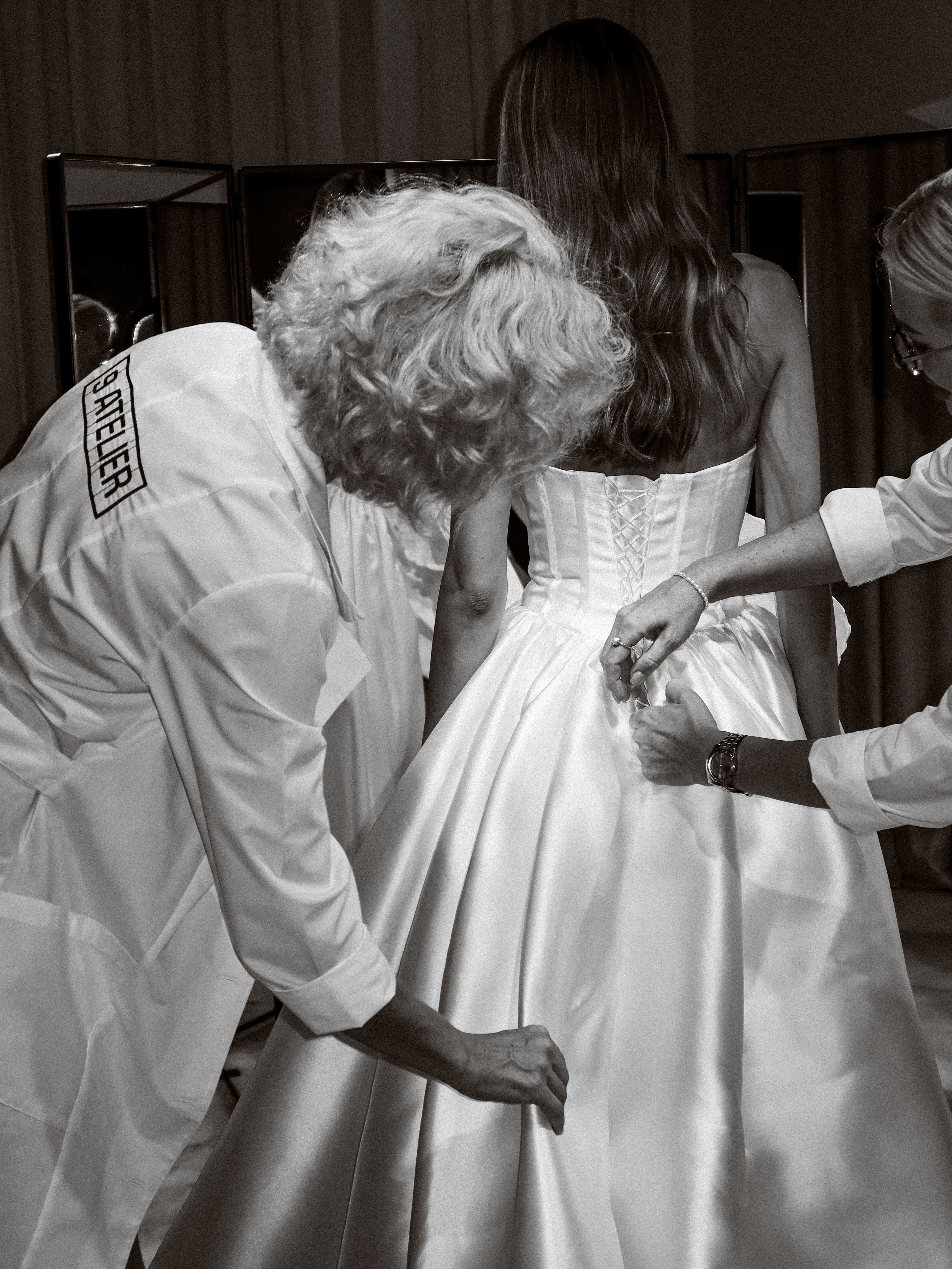 Two women adjusting the back of a bride's satin wedding dress with a corset-style lace-up bodice.