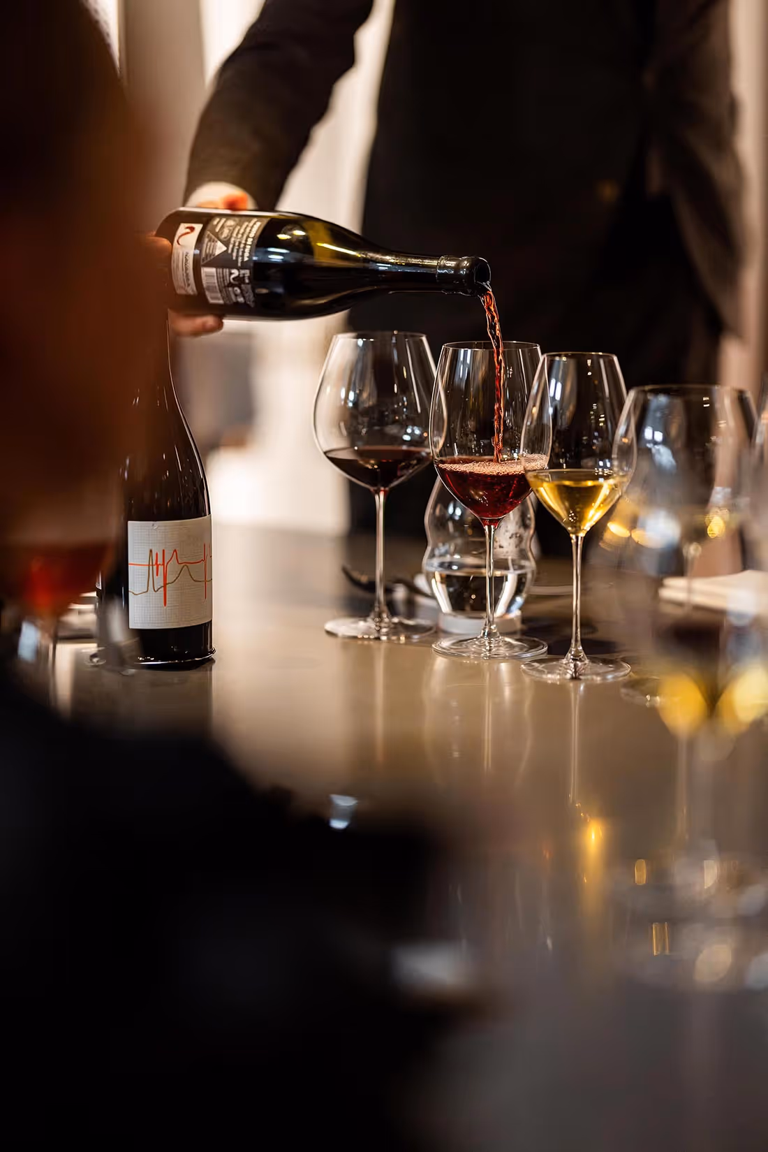 En tjeners hånd hælder rødvin fra en flaske i et glas ved et bord med flere vinglas, der indeholder rød og hvid vin.