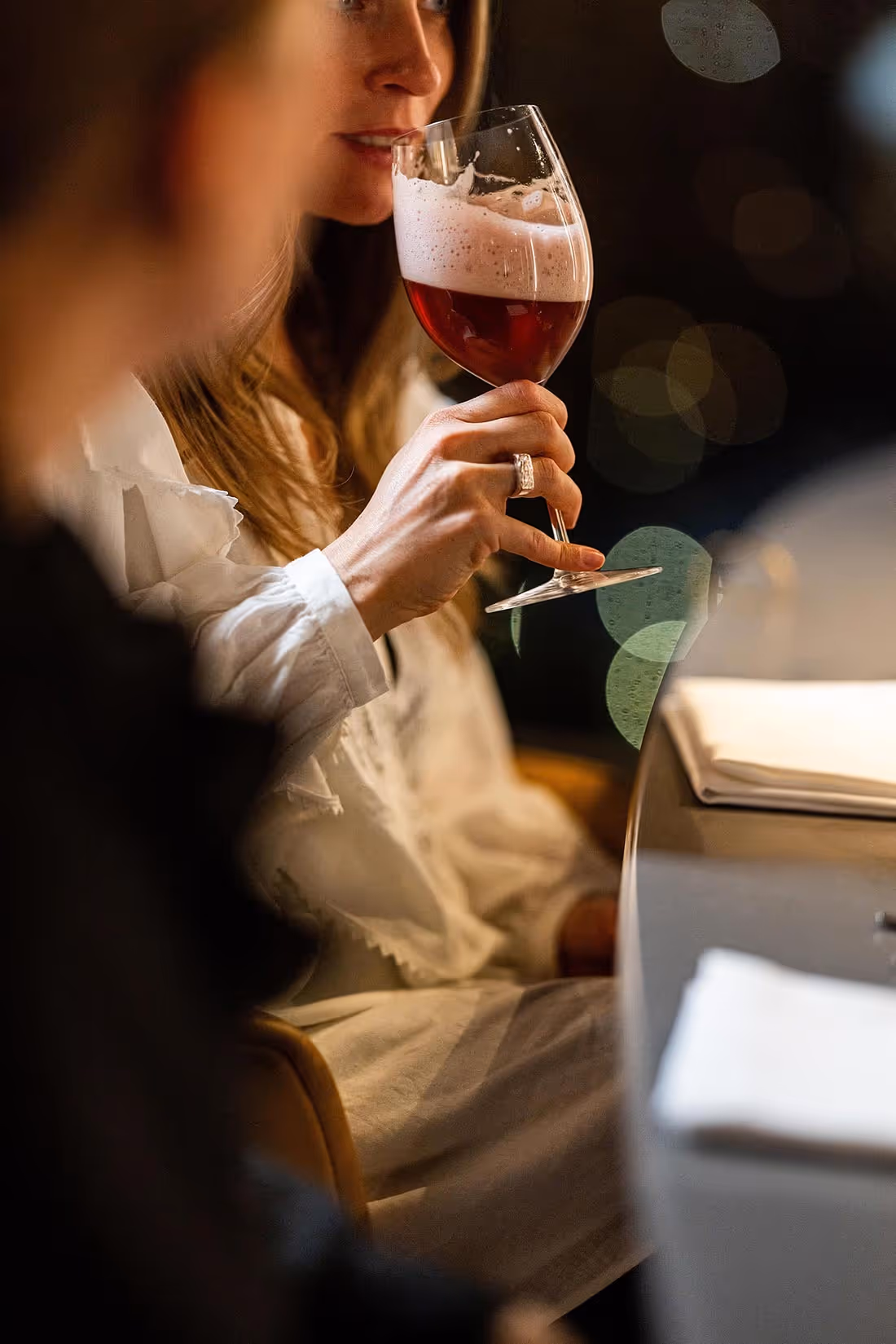 Kvinde i hvid skjorte sidder ved et bord og holder et vinglas med mørk rød saft drik med skum.