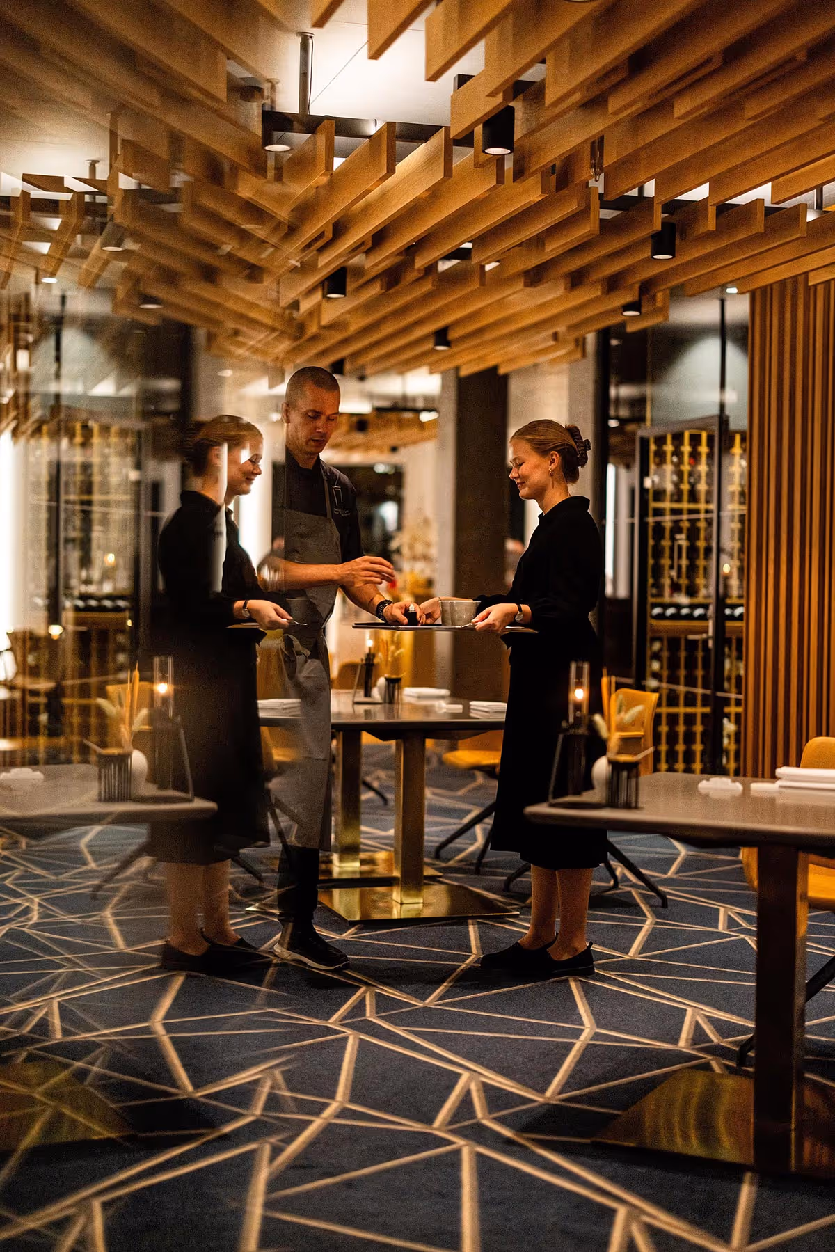 To tjenere udveksler varer på en tallerken i en moderne restaurant med geometrisk tæppe og træloft.