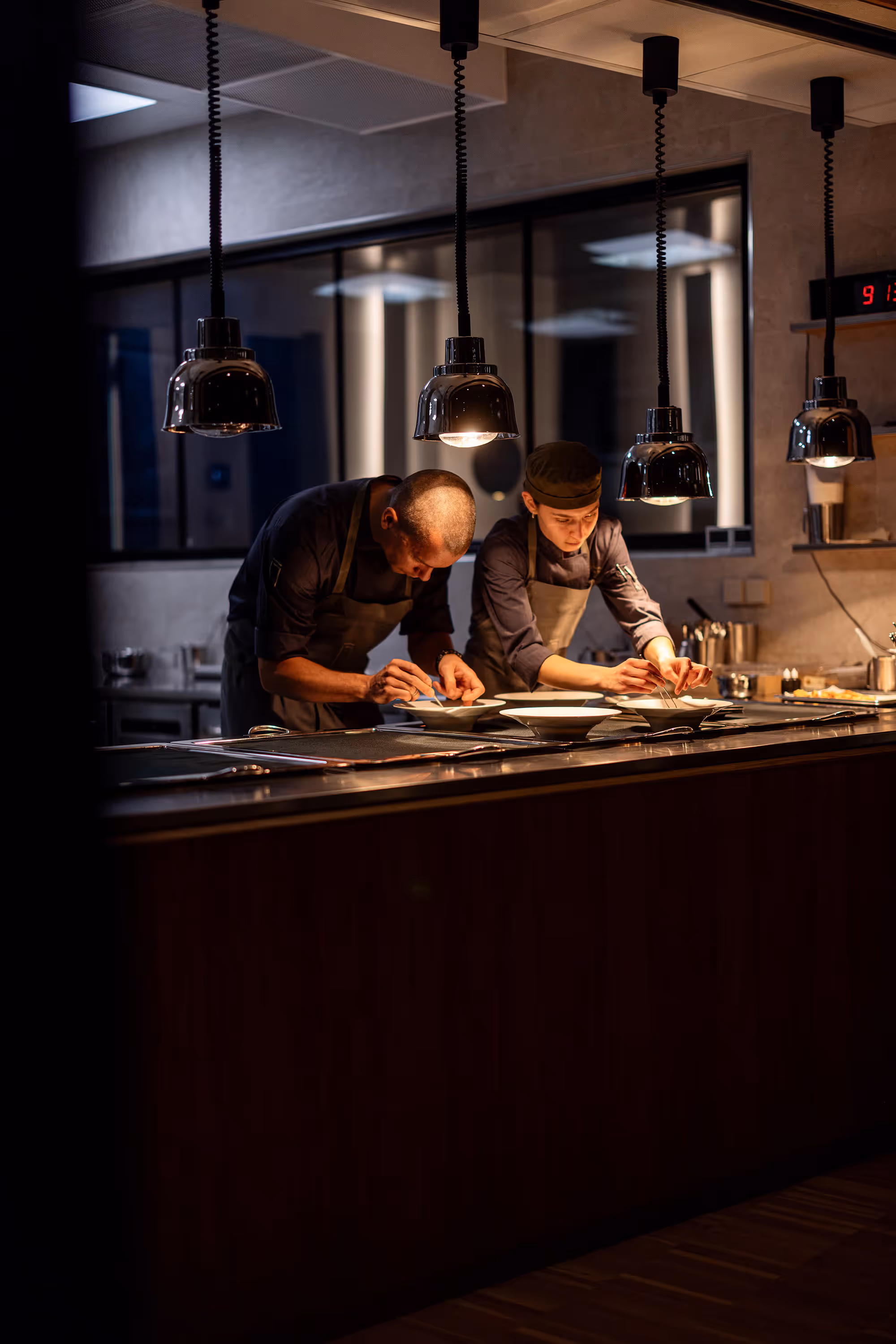 To kokke i mørkt køkken lægger sidste hånd på tallerkener under hængelamper.
