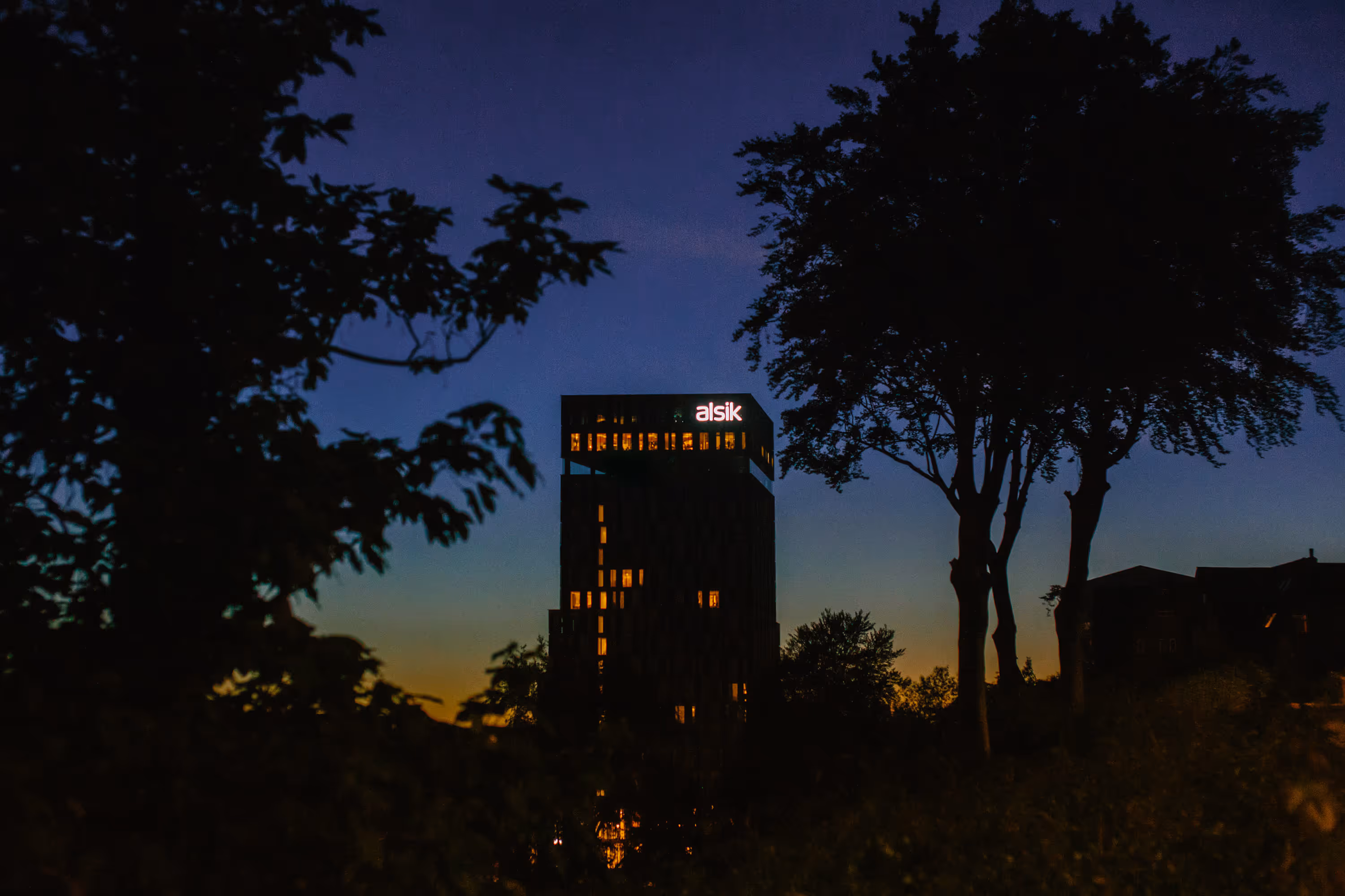 Aftenbillede af en oplyst høj bygning med skiltet 'alsik' omgivet af mørke træer mod en blå-orange himmel.