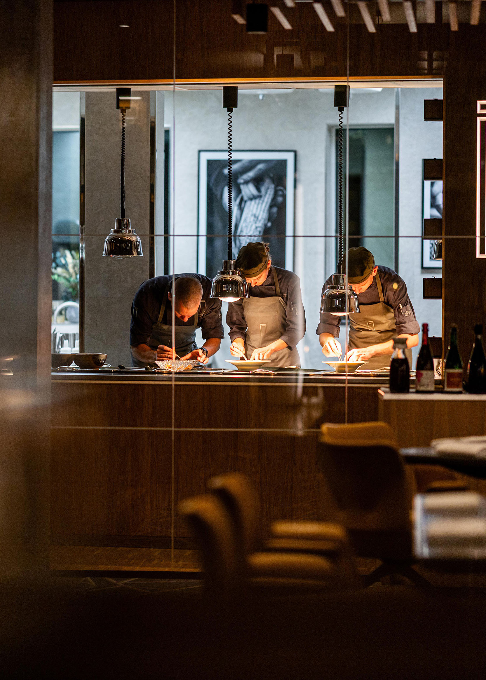 Der ses på afstand tre kokke, heriblandt Peter Rødsgaard, forberede retter på tallerkener.