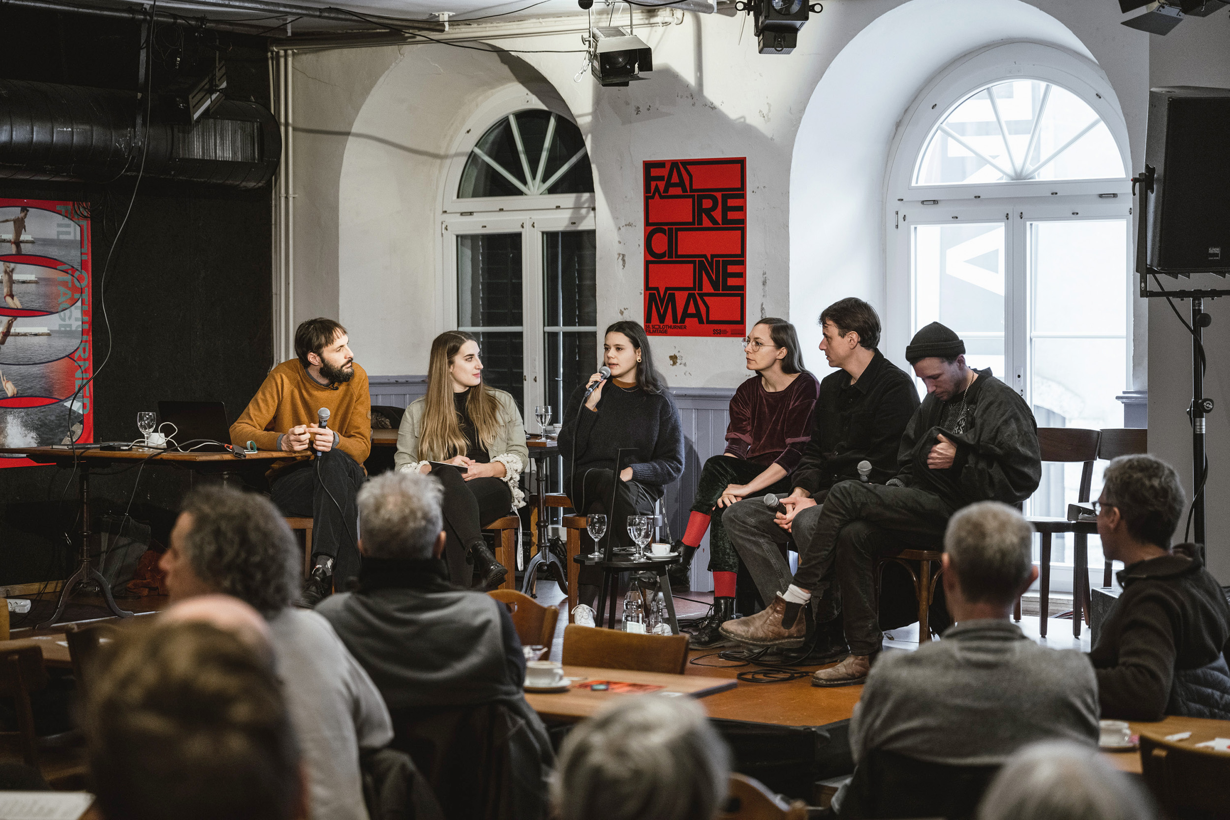 The red "Fare Cinema" visual with black typography in the background of a film discussion with a small podium and a lot of people in front of it