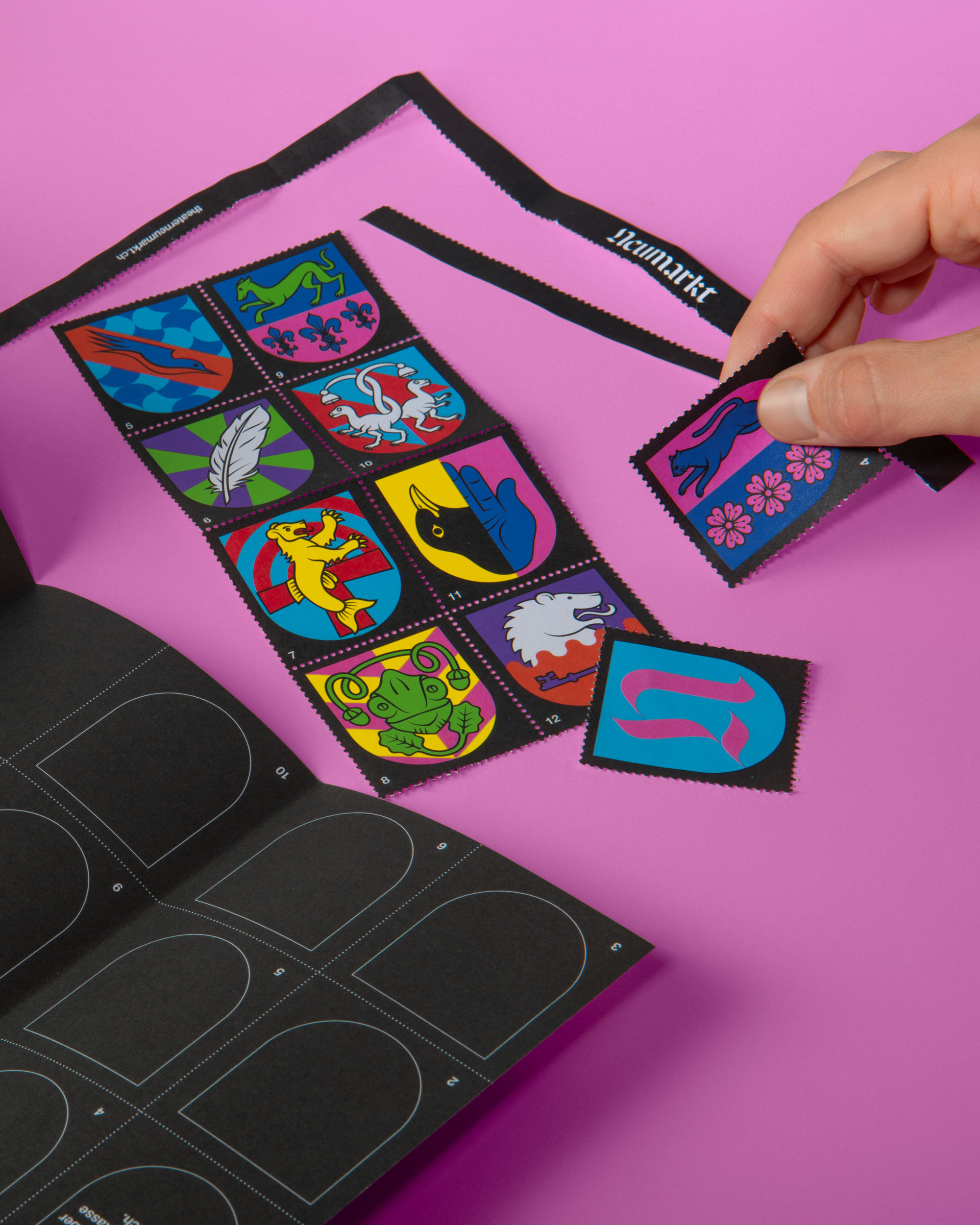 Hand peeling a square colorful sticker featuring a blue cat and pink flowers from a sheet of various vibrant heraldic-style stickers on a pink background.