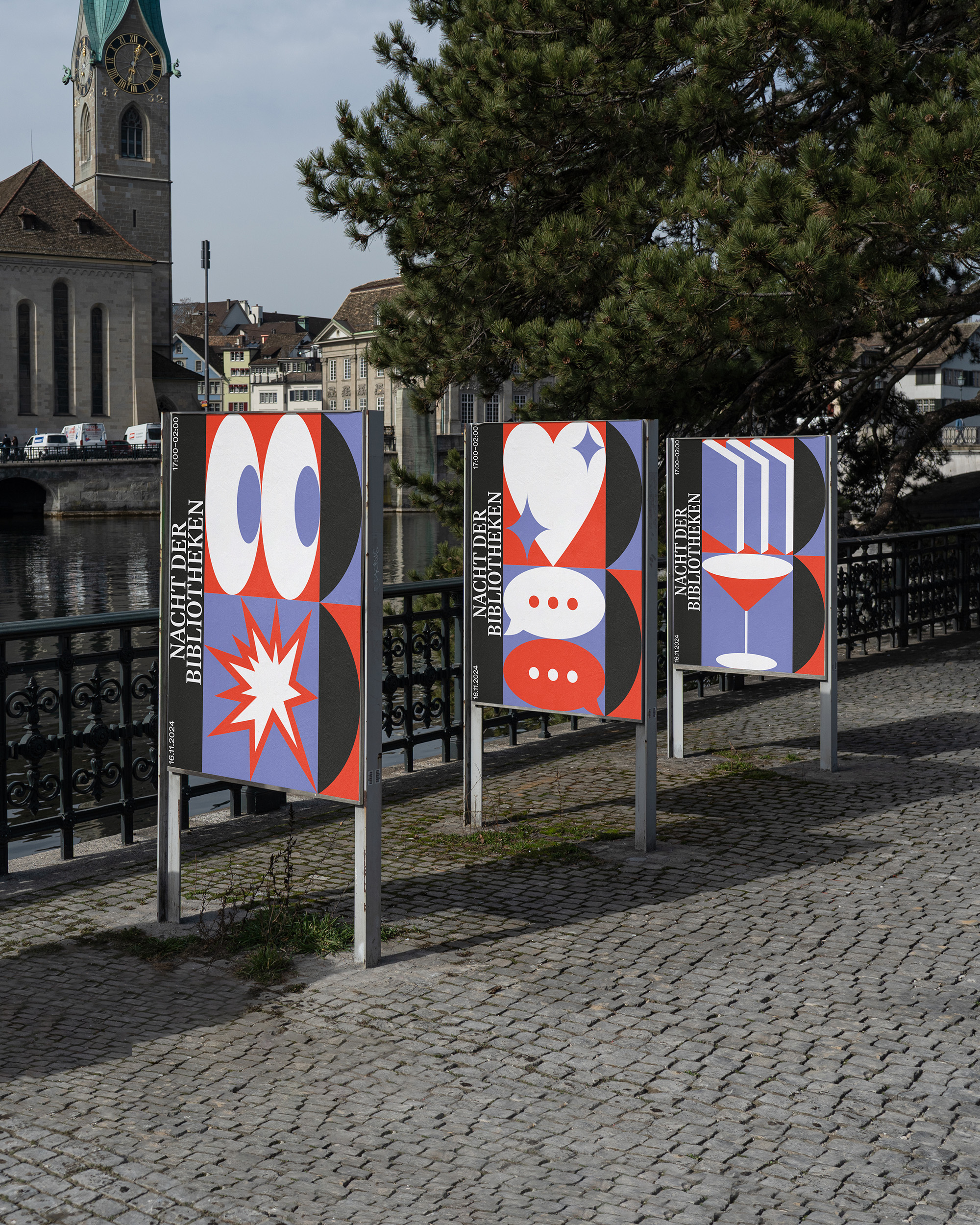 Three colorful signs with abstract shapes and the text 'Nacht der Bibliotheken' along a riverside cobblestone path, with a church tower and buildings in the background.