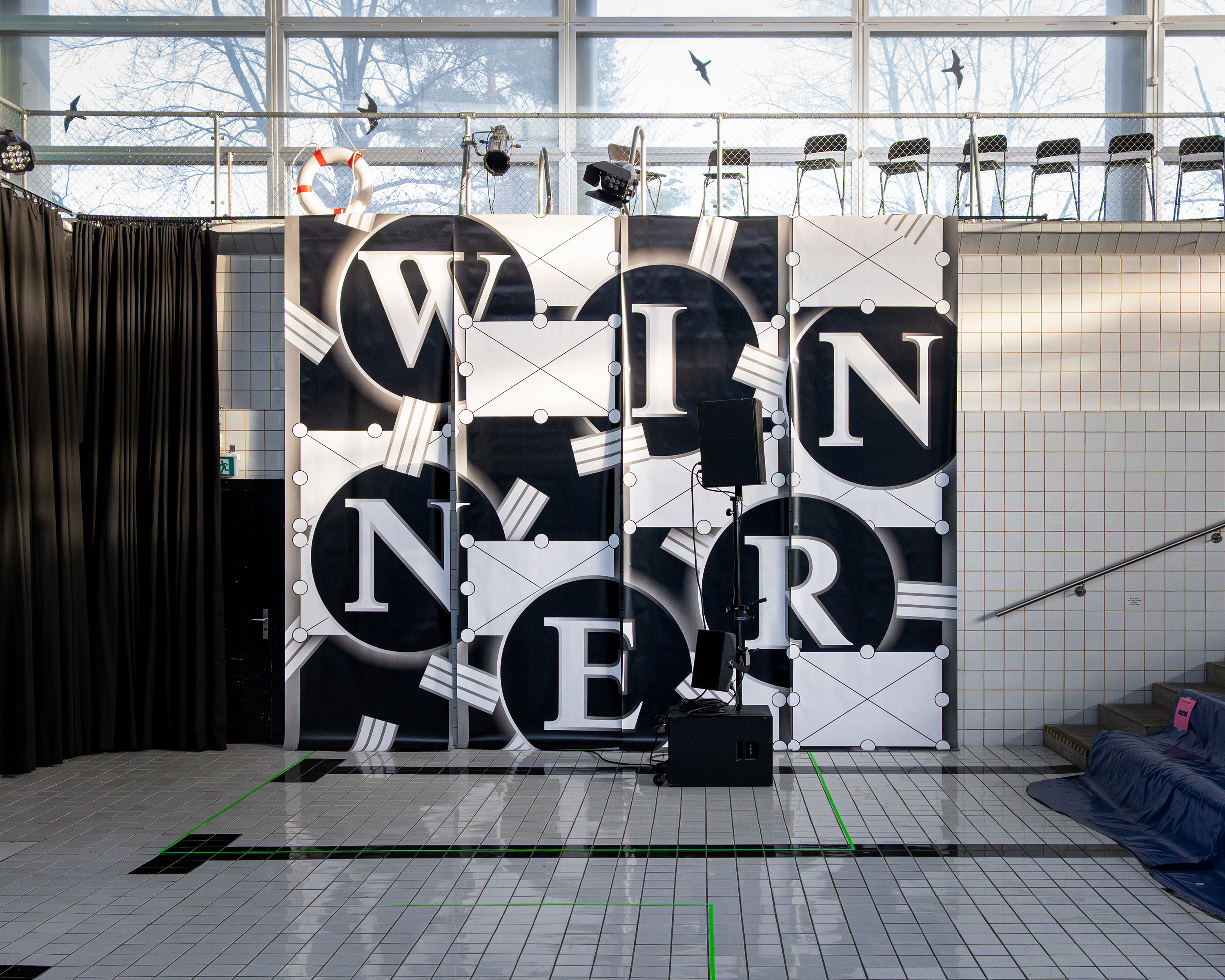 Indoor tiled space with a black and white display spelling 'WINNER' in large letters on panels, with chairs and lighting equipment above.